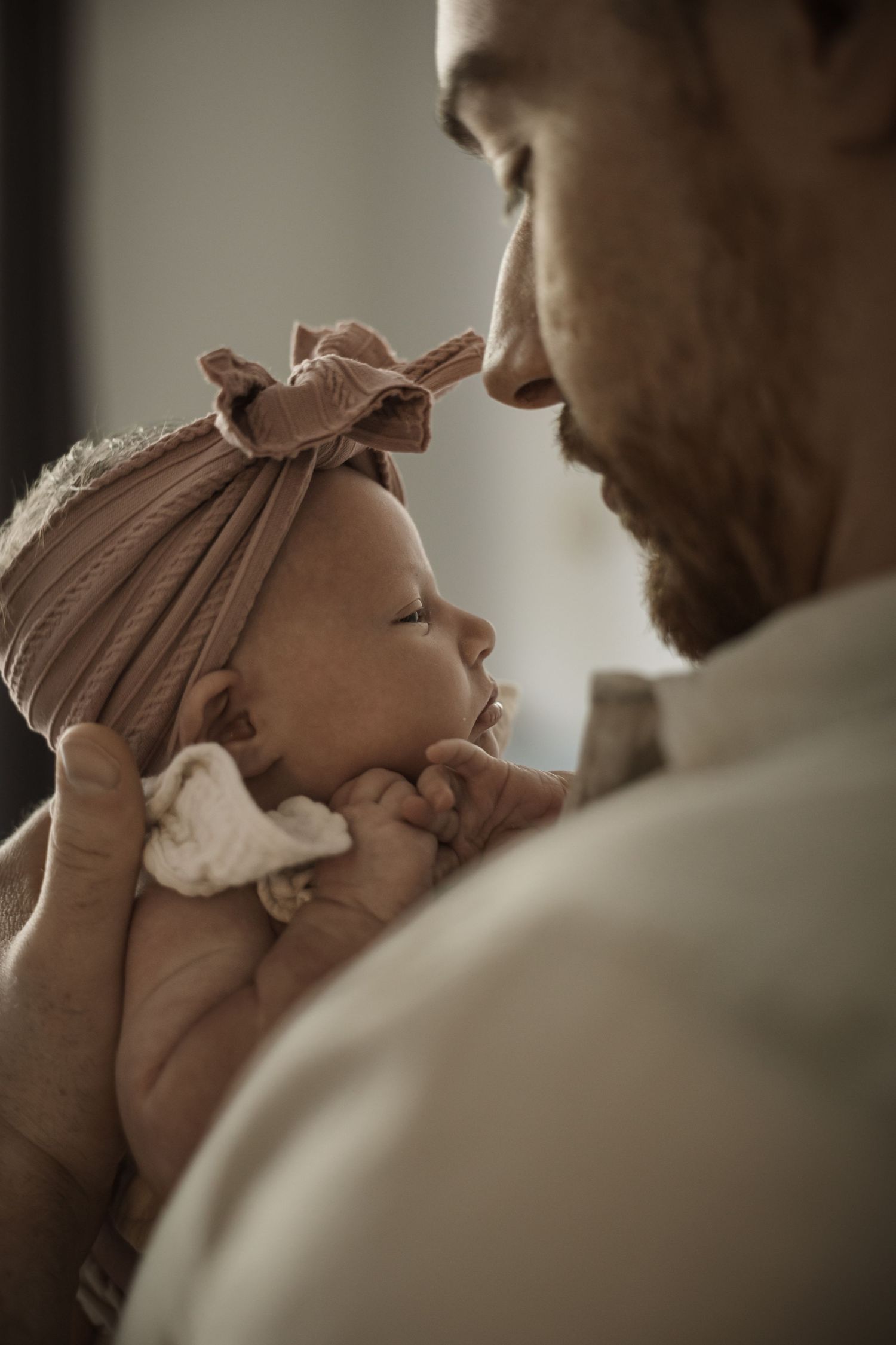 Bearded father holds sleeping newborn baby girl with pink bow headband close to his face.