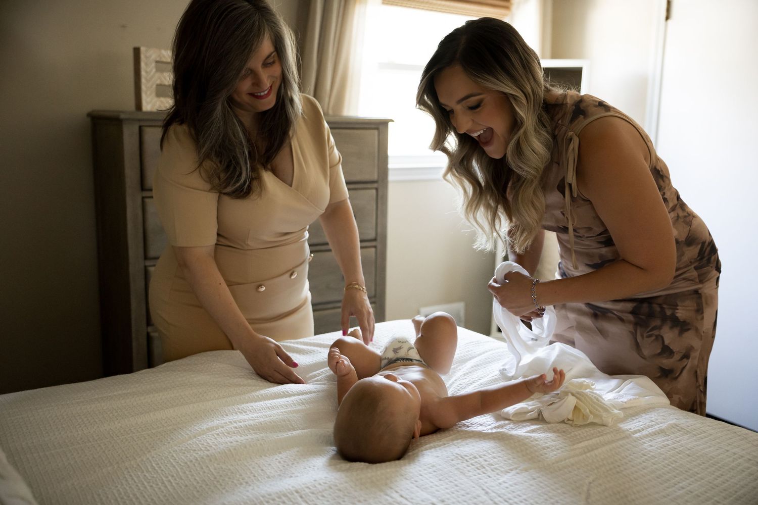 Two women smiling while dressing a newborn baby on a white bed indoors.