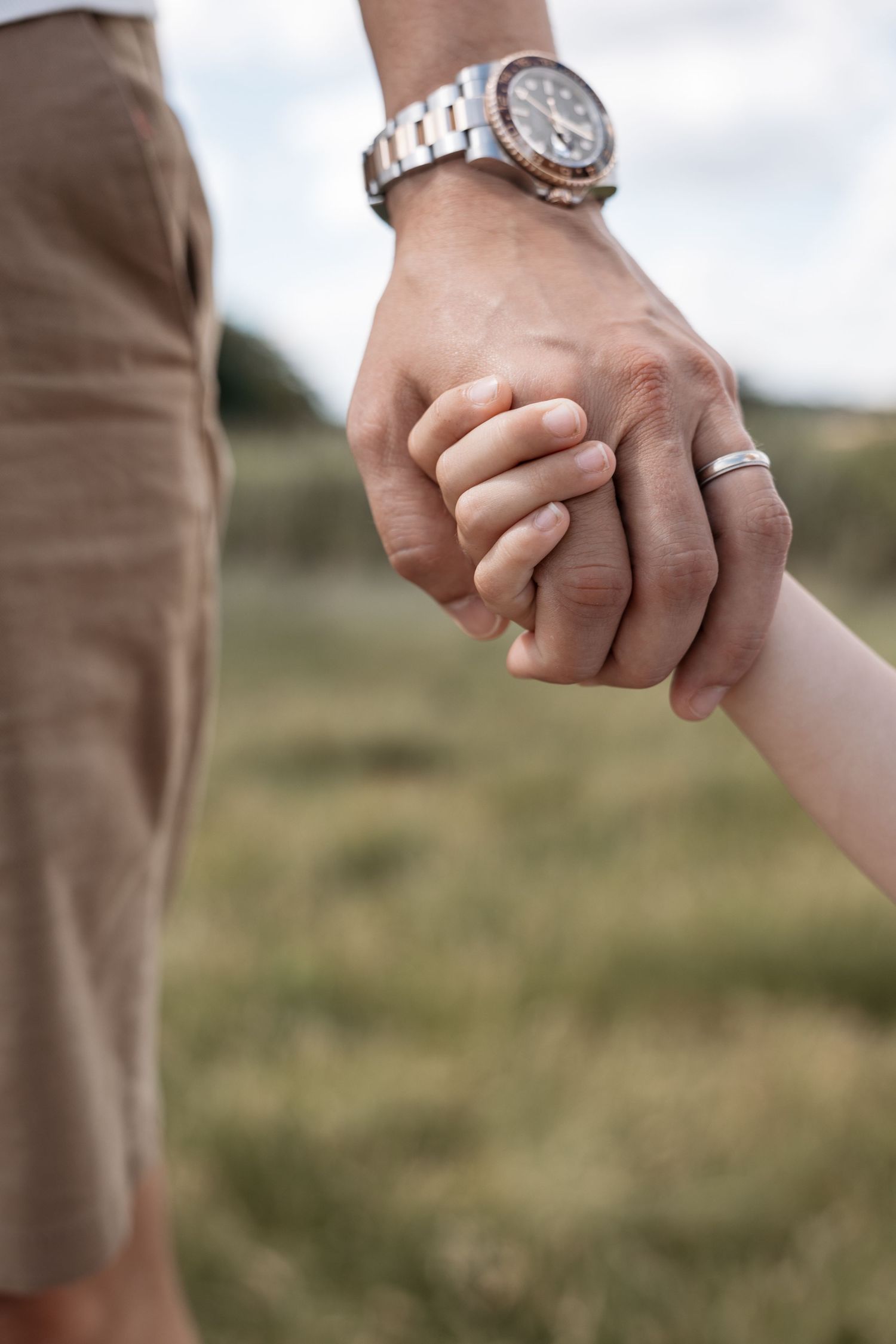 A close-up of an adult hand wearing a watch and ring gently holding a child's hand.
