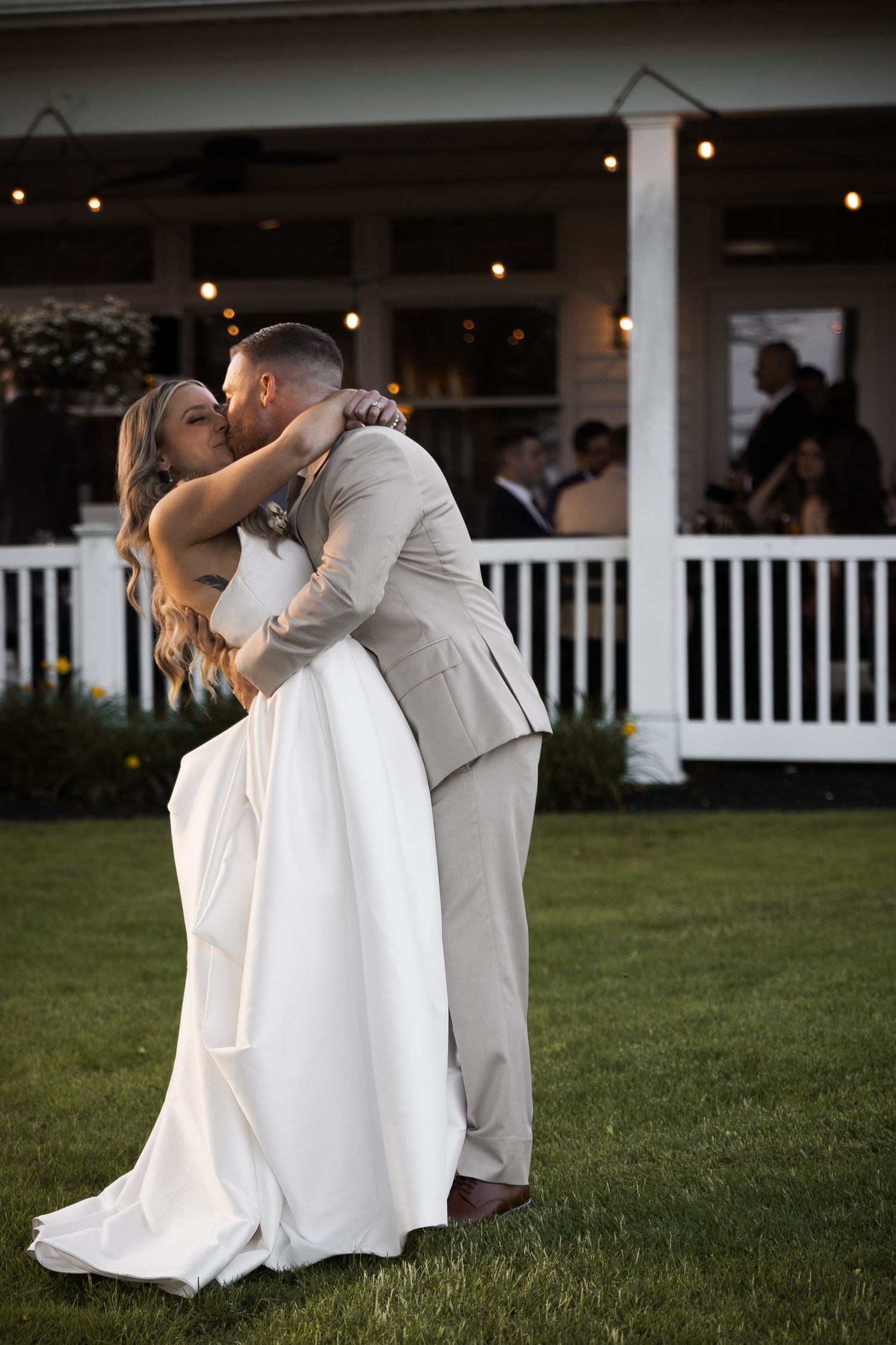 Groom lifts bride in white wedding gown for a kiss on lawn outside white porch venue at golden hour in New Jersey.