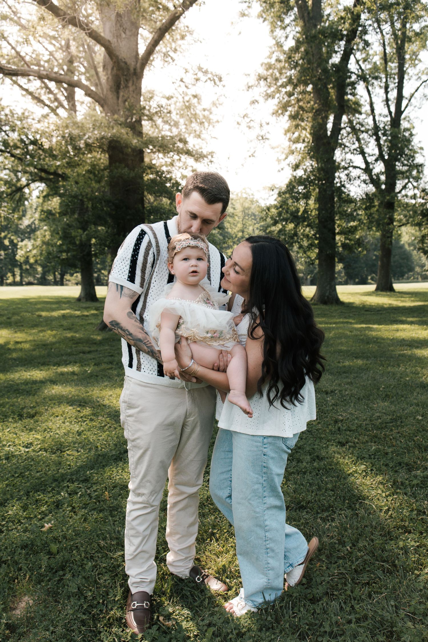 Young family with tattooed dad and dark-haired mom holding baby girl in white dress at sunny park.