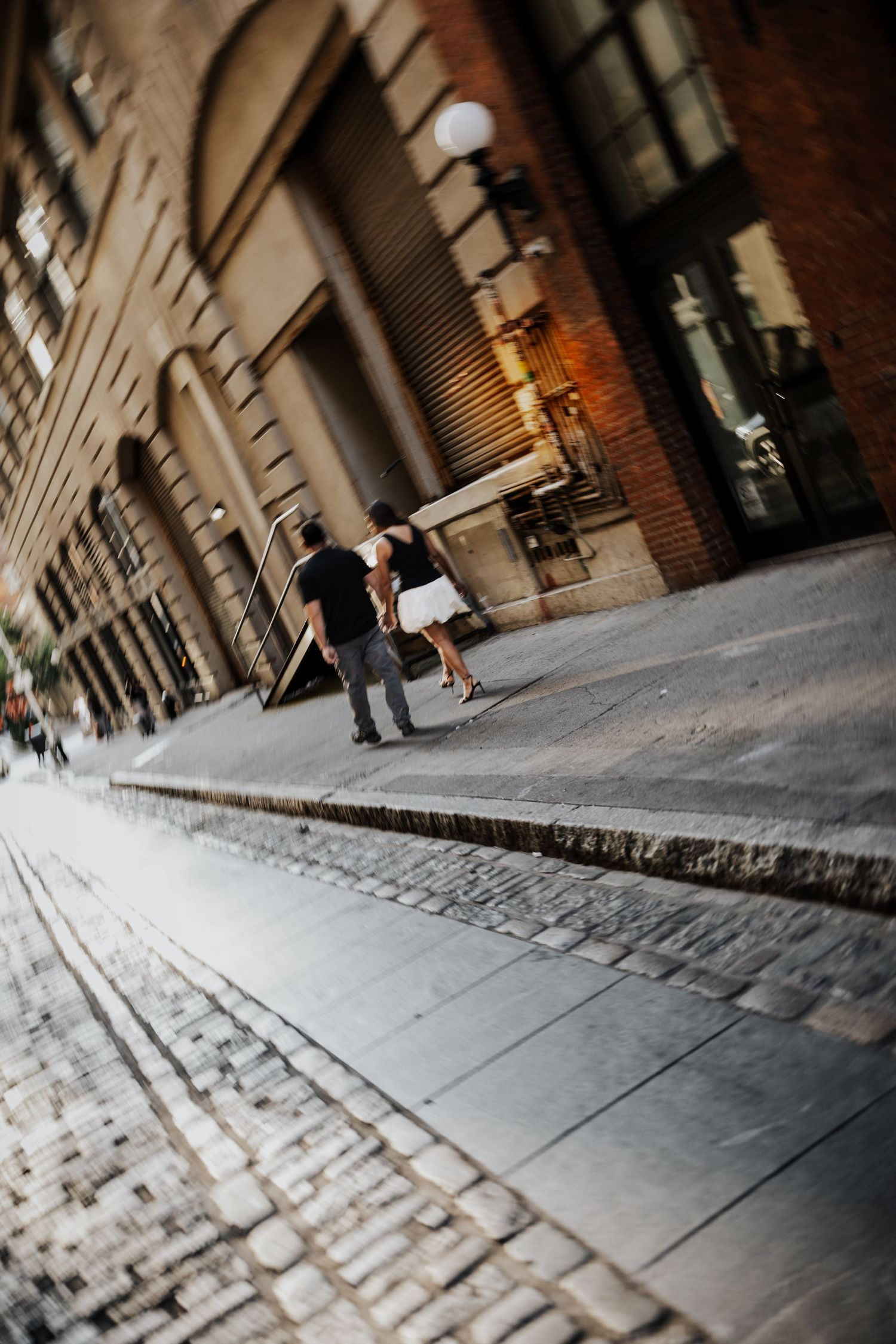 Couple walking along cobblestone street beside historic urban building during engagement session.