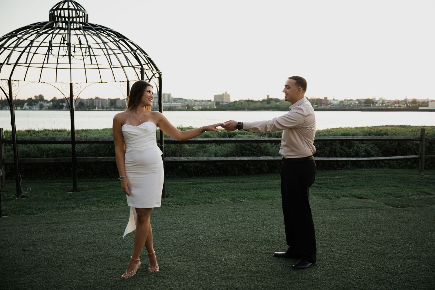 Couple holding hands near an ornate gazebo with a waterfront view at dusk.