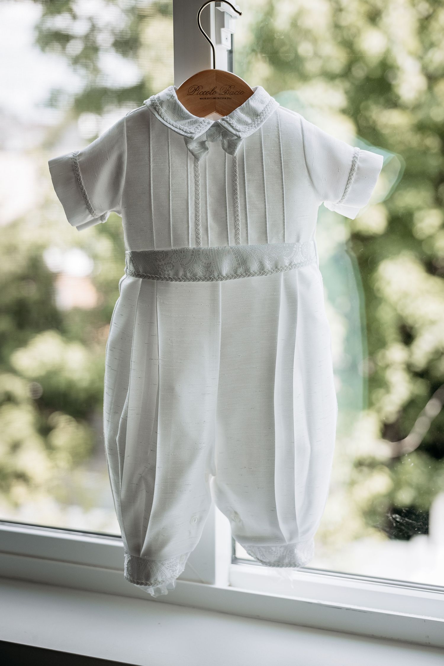 White baby christening romper suit hanging on wooden hanger by window.
