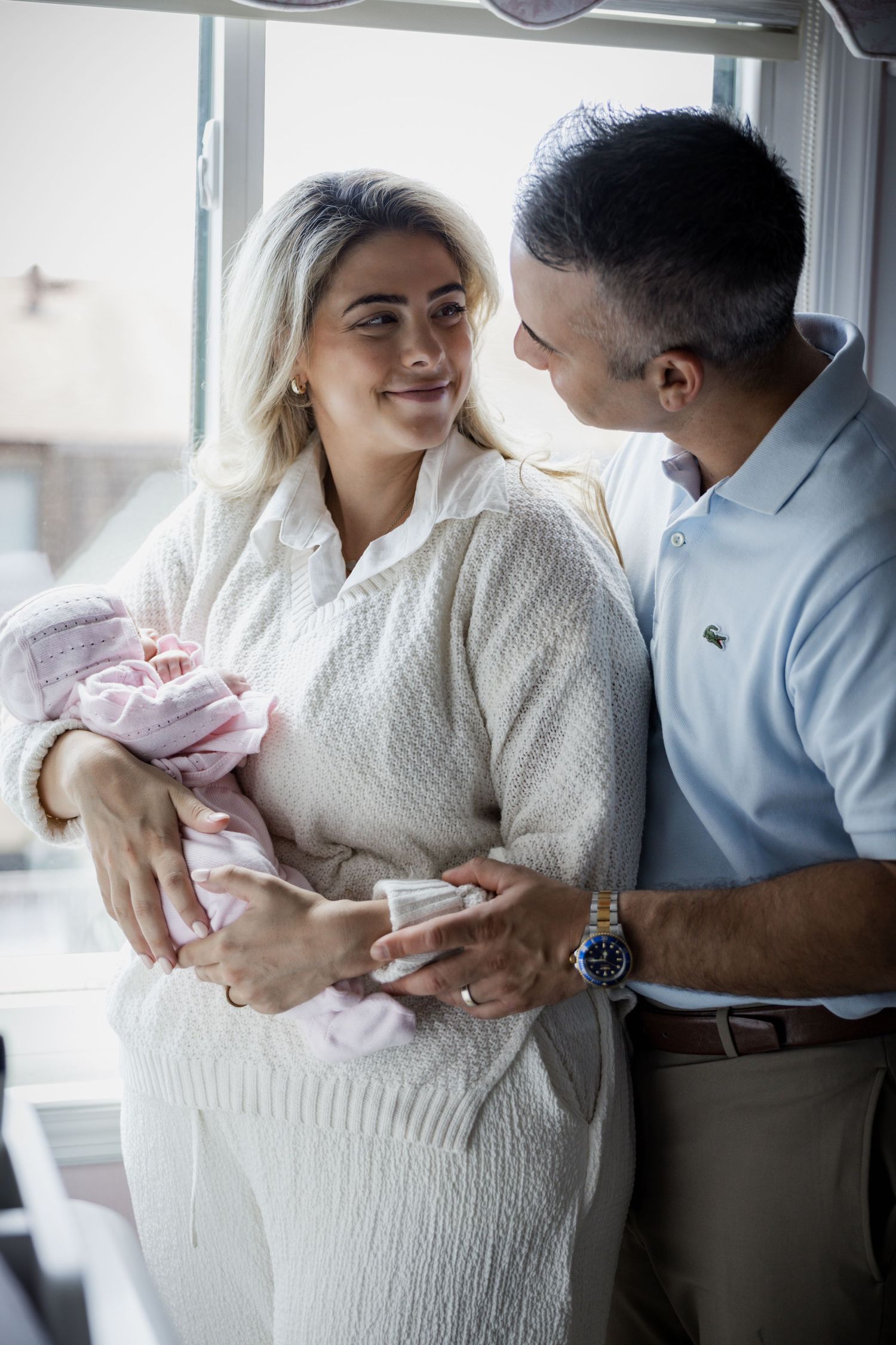 Smiling mother holds newborn in pink while father looks on lovingly by window.