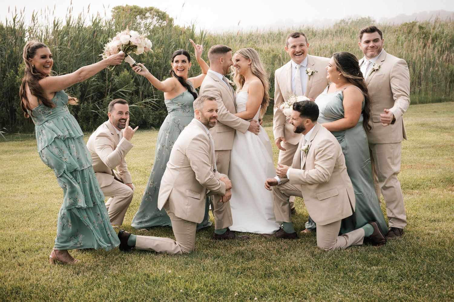 A joyful wedding party in sage green and tan attire celebrates together on a lush green lawn in New Jersey.
