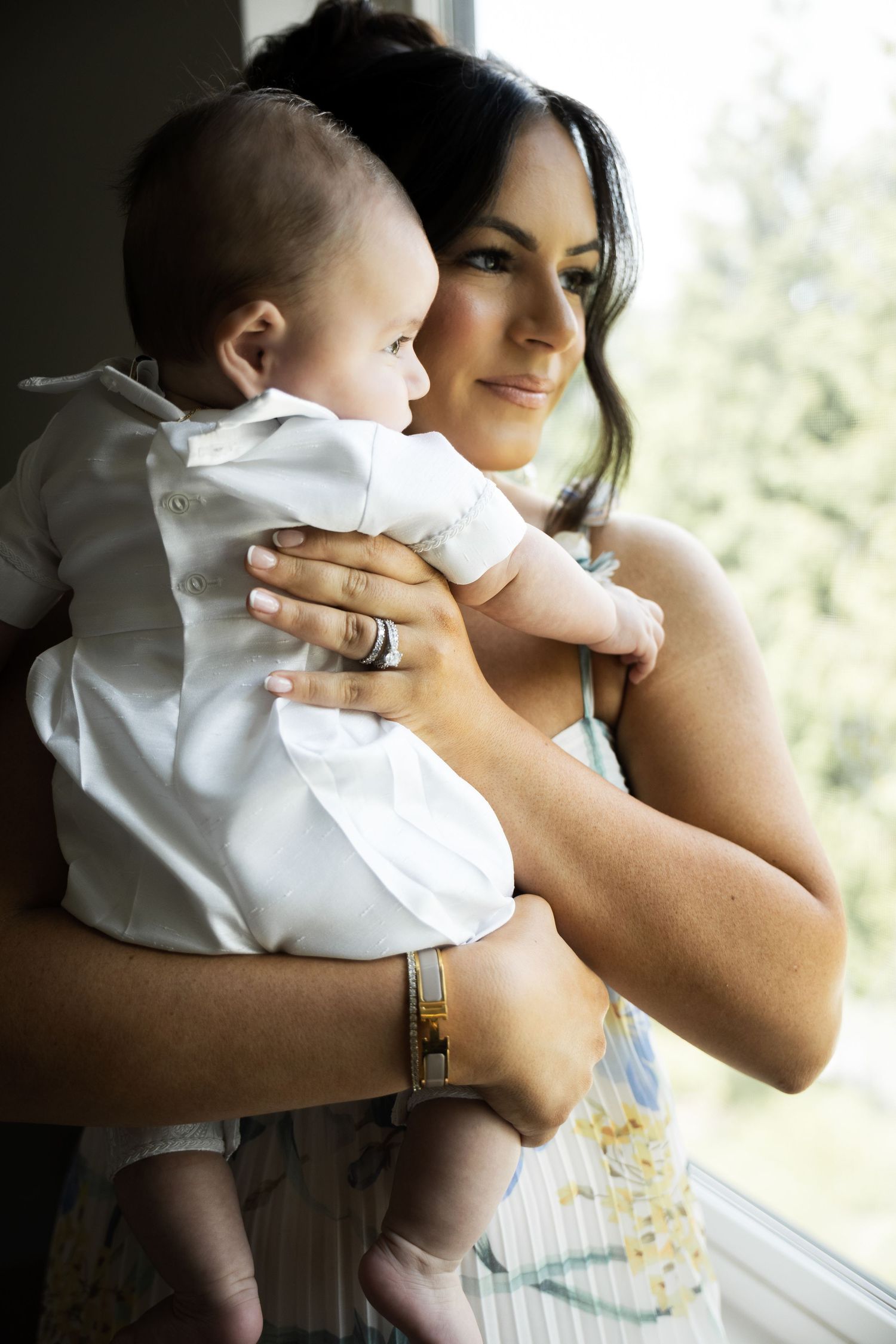 Dark-haired mother tenderly holding baby in white christening outfit by window in Staten Island.