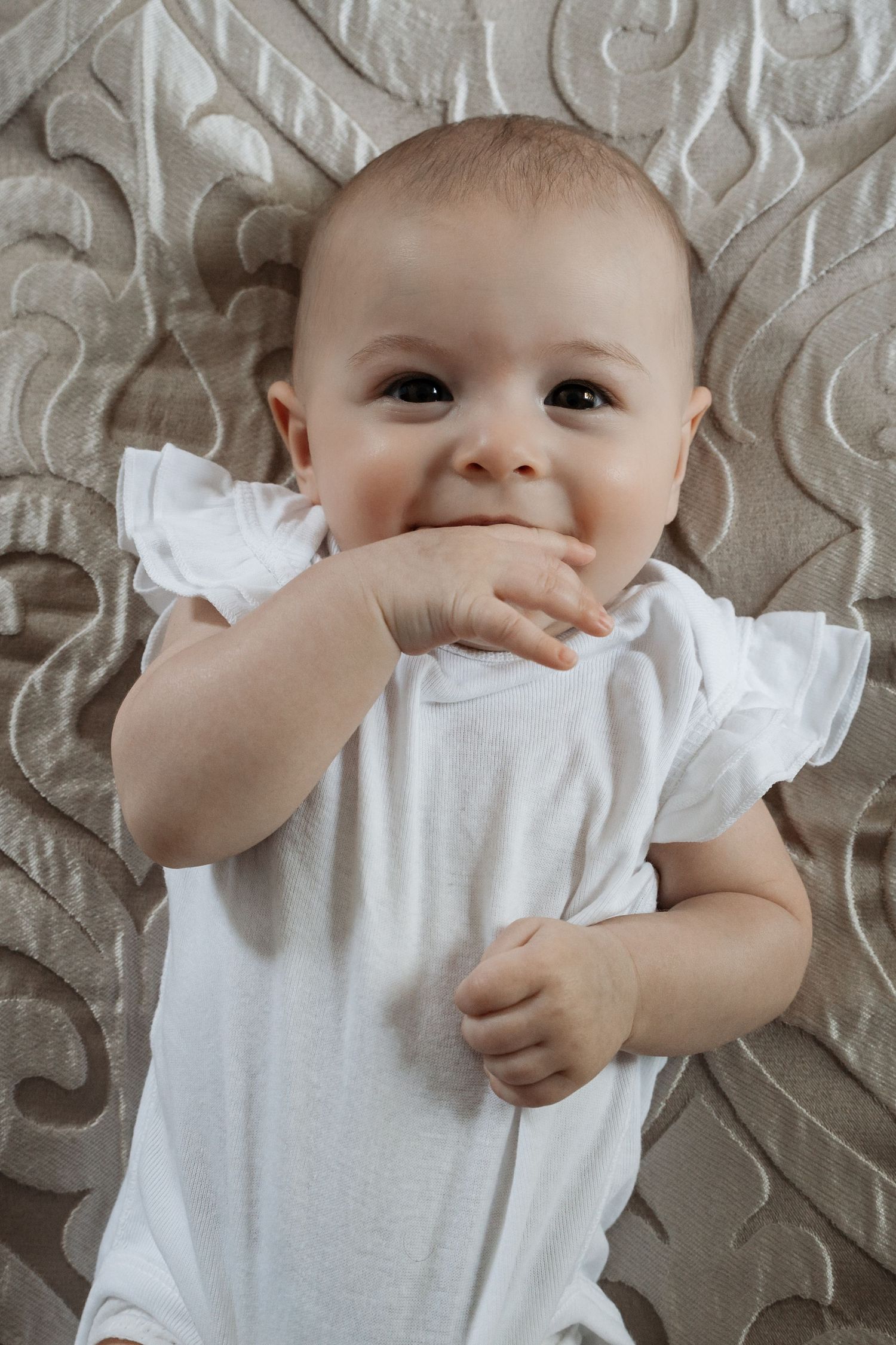 Adorable baby in white onesie lying on textured beige surface with hand near mouth smiling.