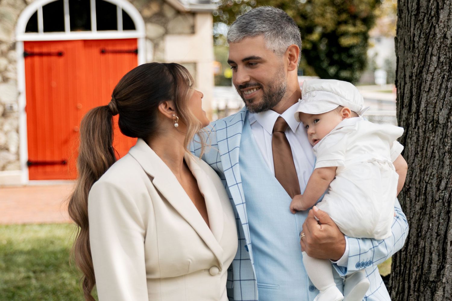Elegant couple in formal attire smiling at baby in white christening outfit outdoors.