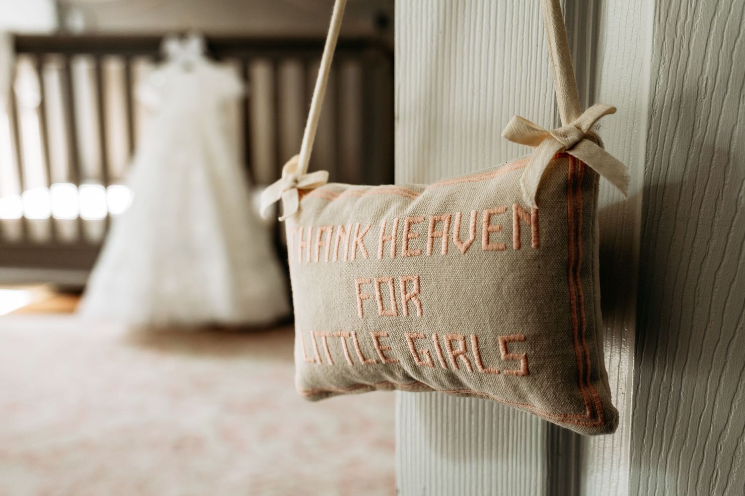 Decorative burlap pillow reading Thank Heaven For Little Girls hanging in nursery with christening gown background.