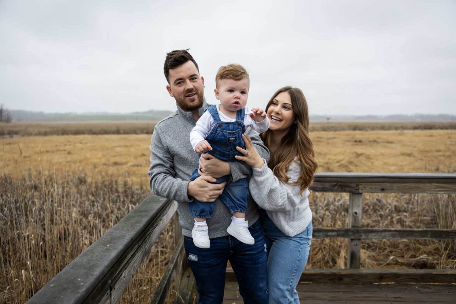 Happy young family with baby boy posing on rustic wooden bridge overlooking golden marsh field.