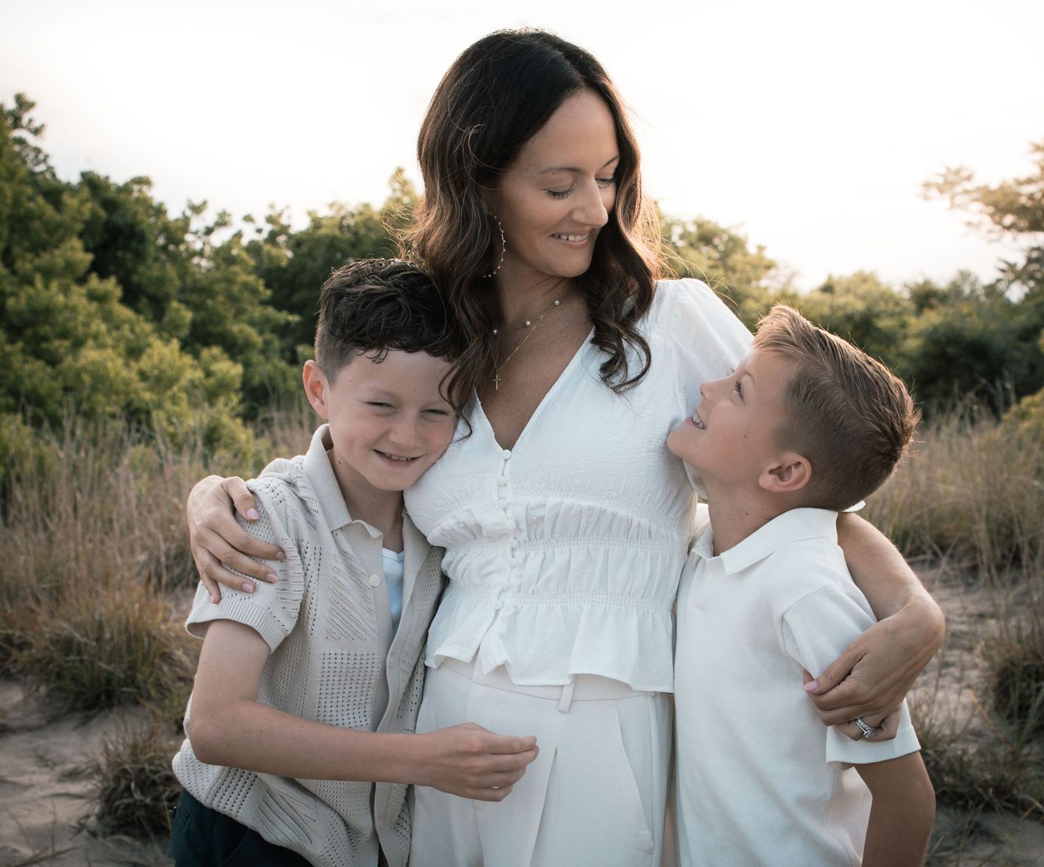 A pregnant mother smiles warmly while embracing her two young sons outdoors in a field.