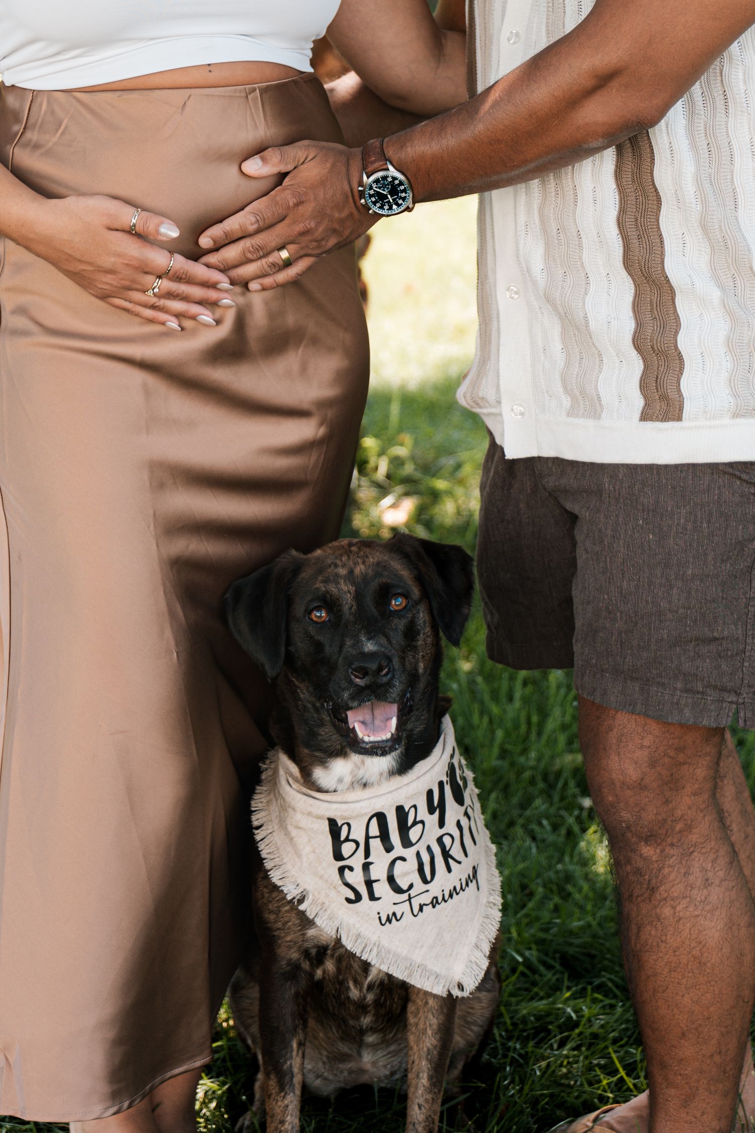 Dog wearing baby security bandana sits between expectant couple outdoors.