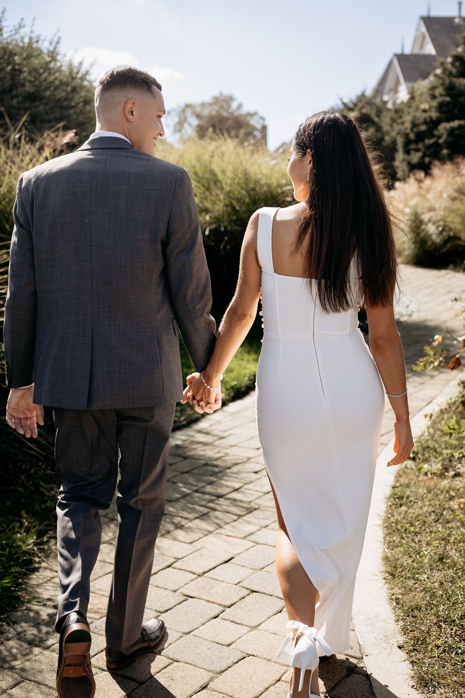 Bride and groom holding hands walking down a sunny brick path in a garden setting.