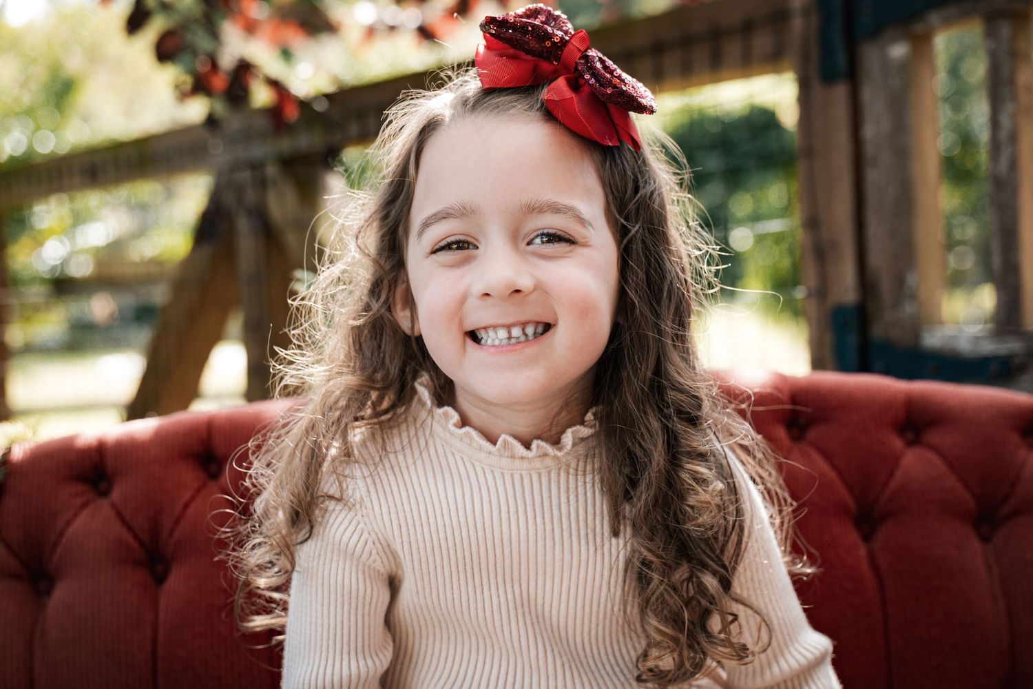 A smiling young girl with curly hair and a red bow sits on a red velvet chair for a holiday portrait.