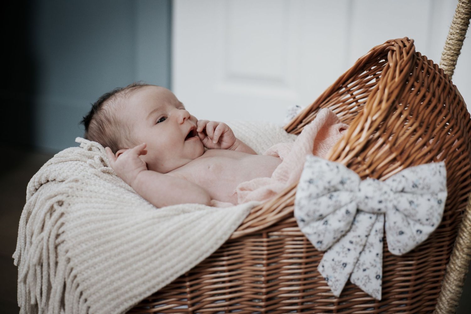 Smiling newborn baby lying in wicker bassinet with cream blanket and floral bow decoration.