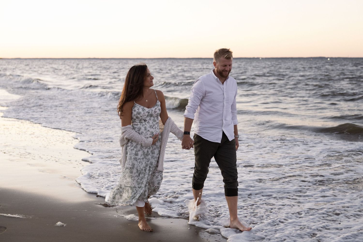A barefoot couple walks hand-in-hand along a serene beach shore at sunset in casual elegant attire in New Jersey.