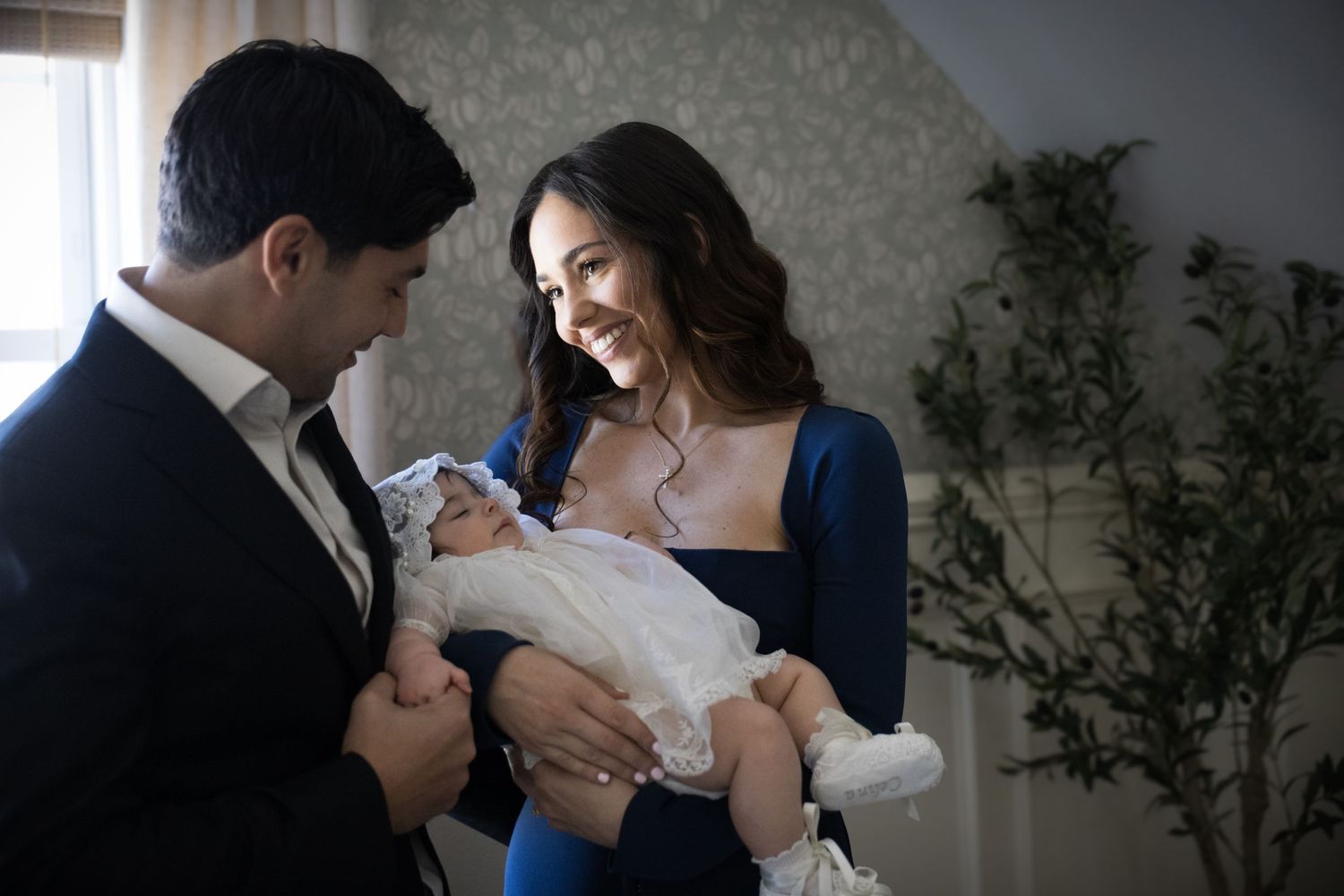 Smiling couple in formal attire tenderly holding their newborn dressed in a white christening gown.