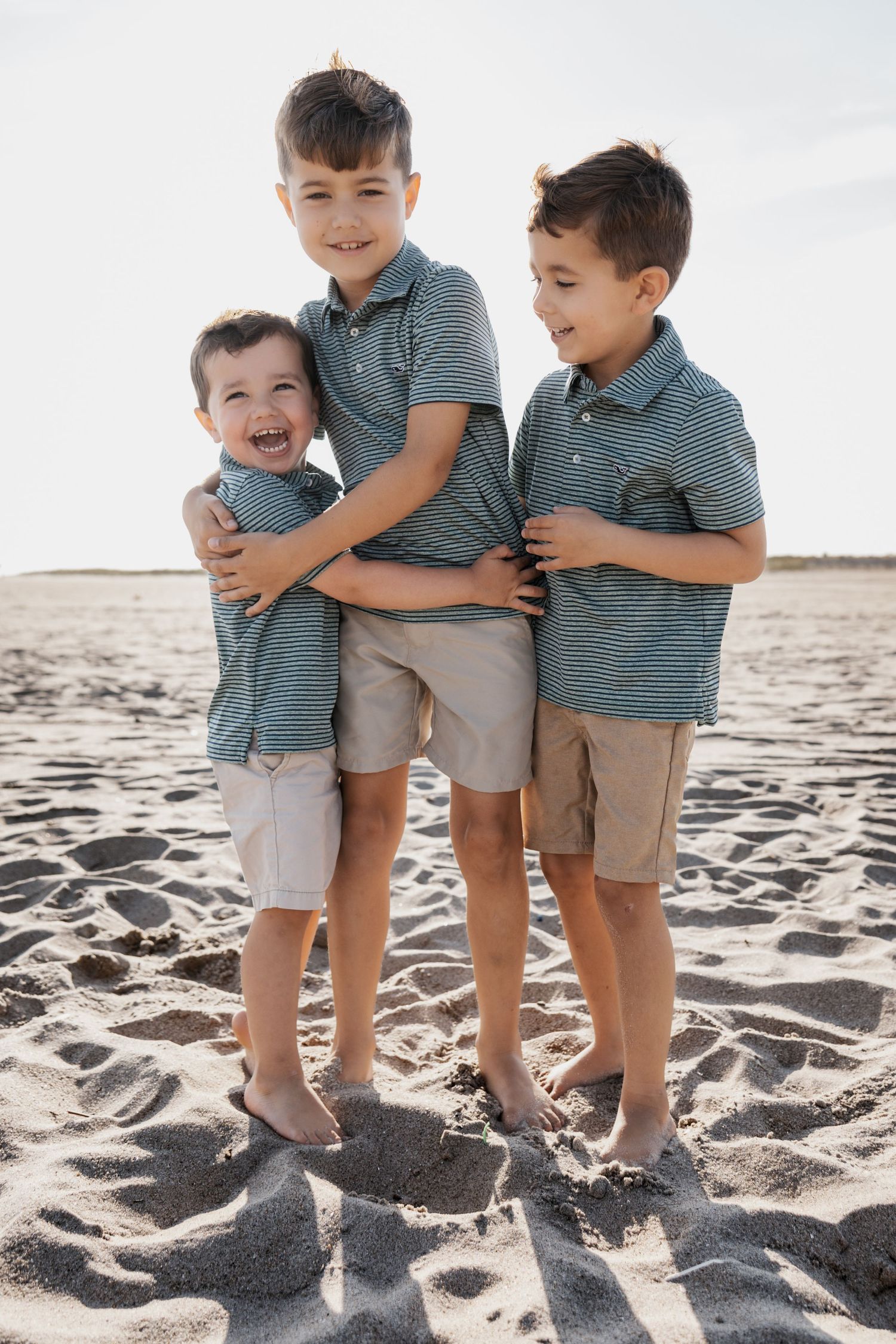 Three brothers in matching striped polo shirts hugging and laughing together on a sandy beach.
