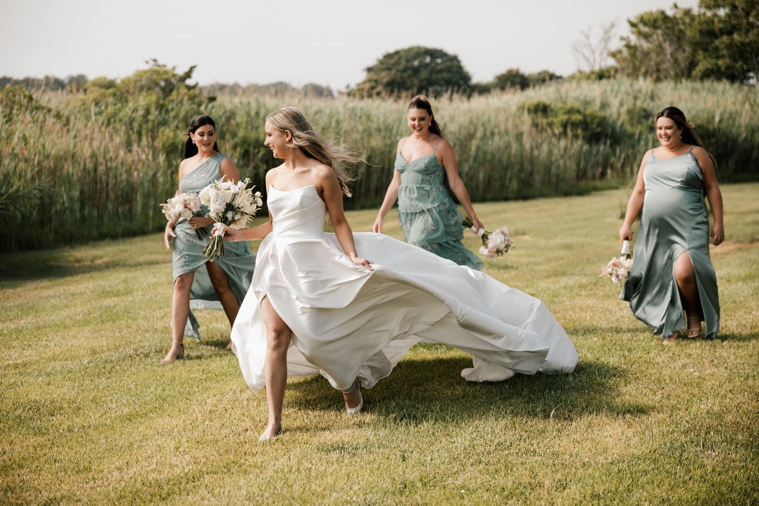 A radiant bride lifts her white gown while walking with bridesmaids in sage green dresses outdoors in New Jersey.