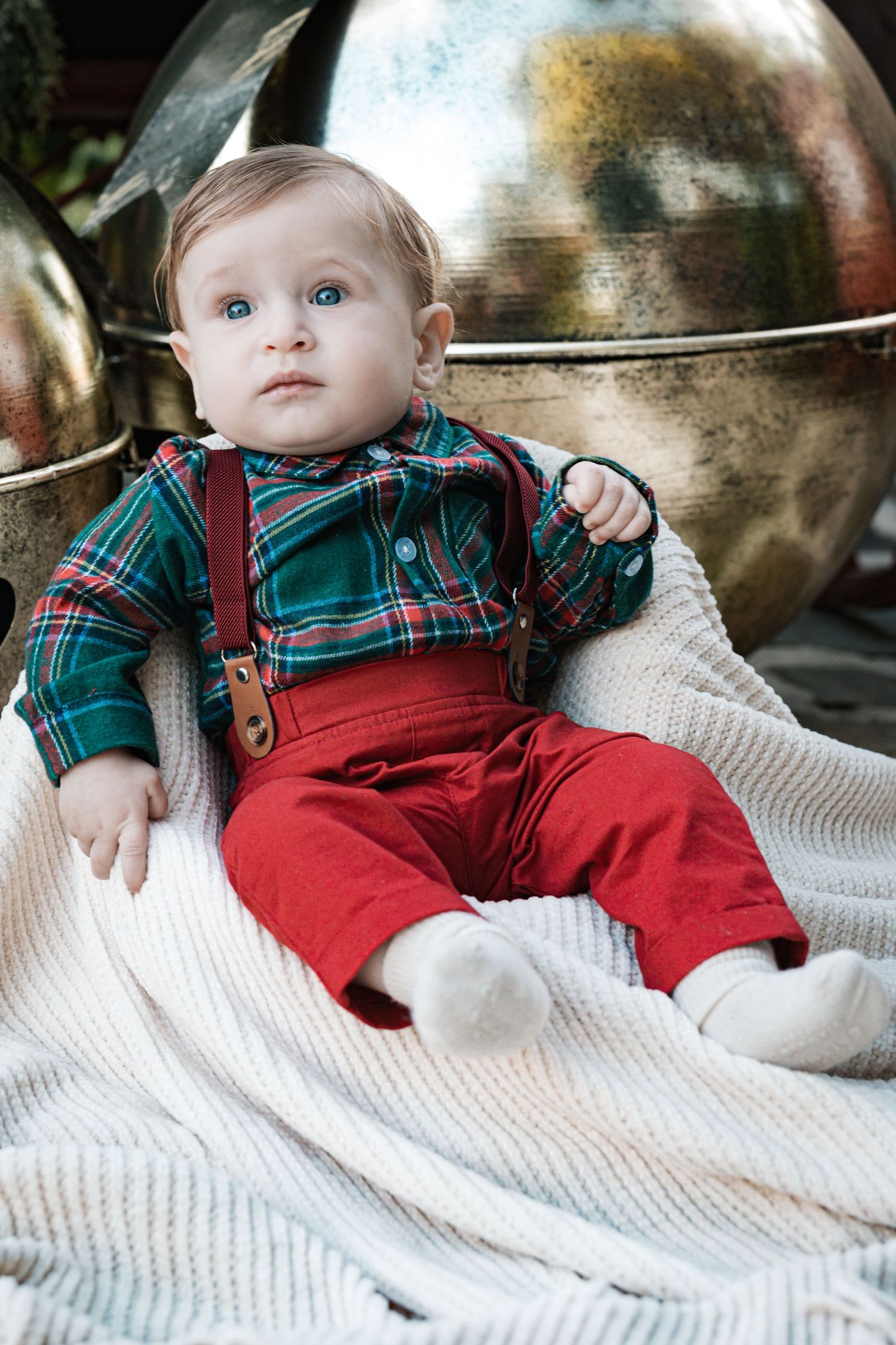 An adorable baby boy in a green plaid shirt and red pants with suspenders sits on a white blanket.
