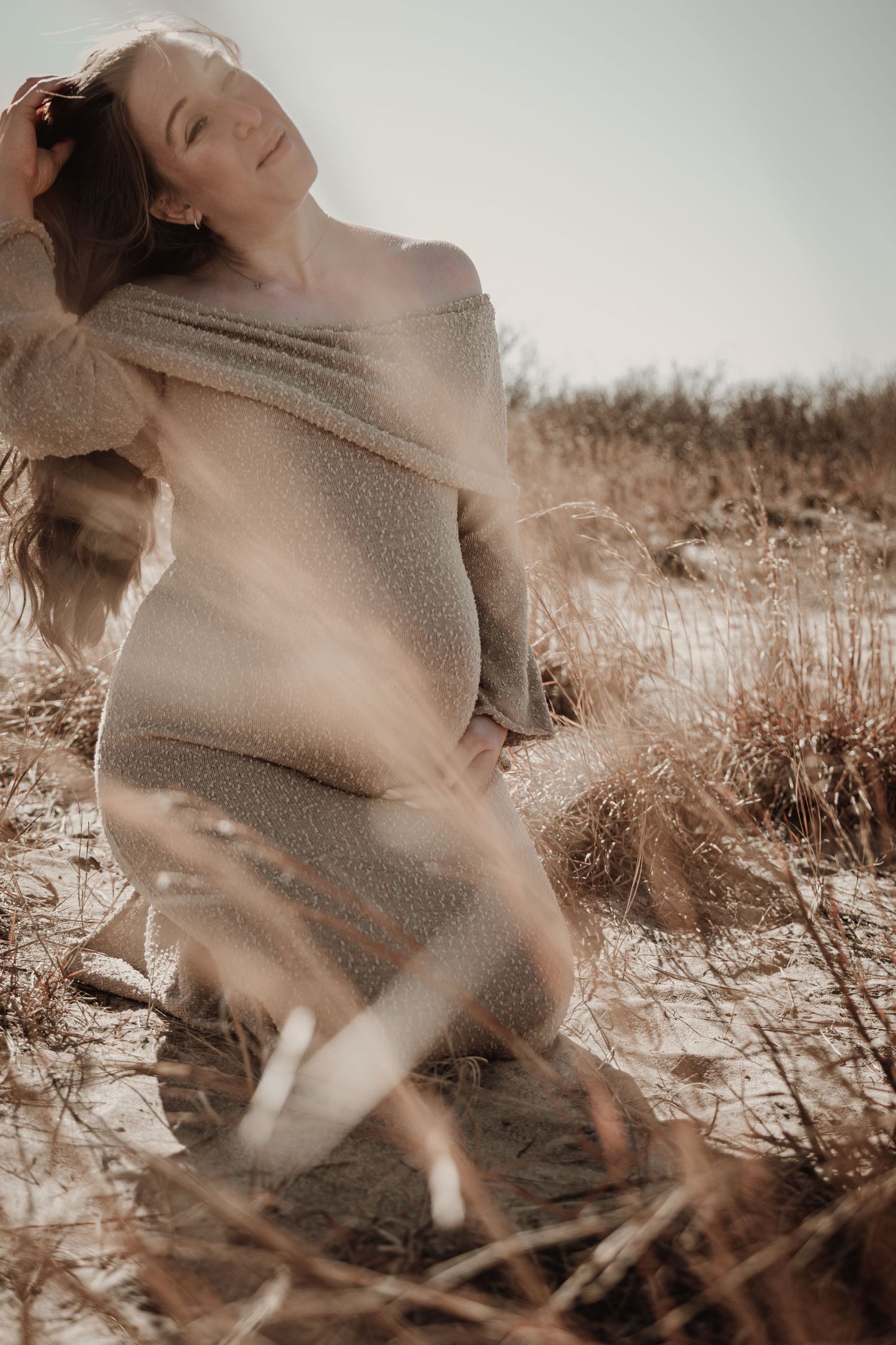 Pregnant woman in off-shoulder beige knit dress kneeling in sandy field during golden hour shoot.