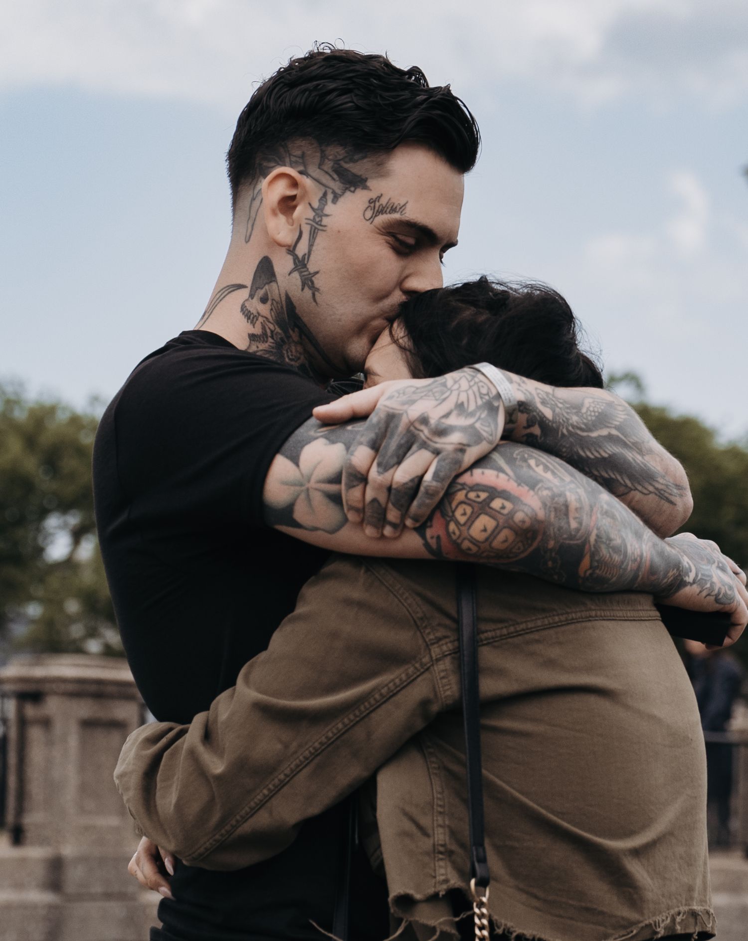 Heavily tattooed couple sharing an emotional embrace outdoors with cloudy sky behind them in New Jersey.