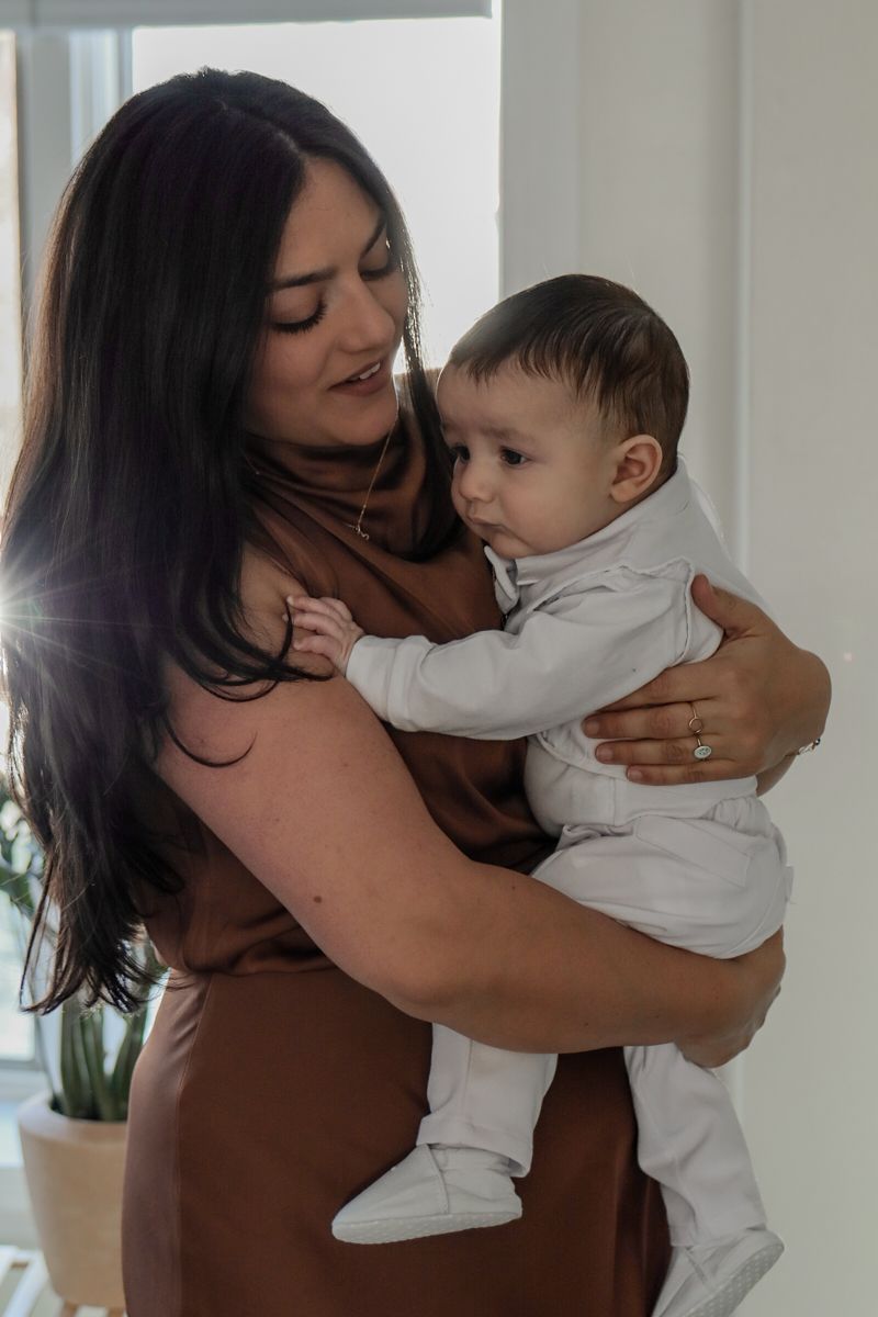 Mother in brown dress tenderly holding her baby boy near a bright window.