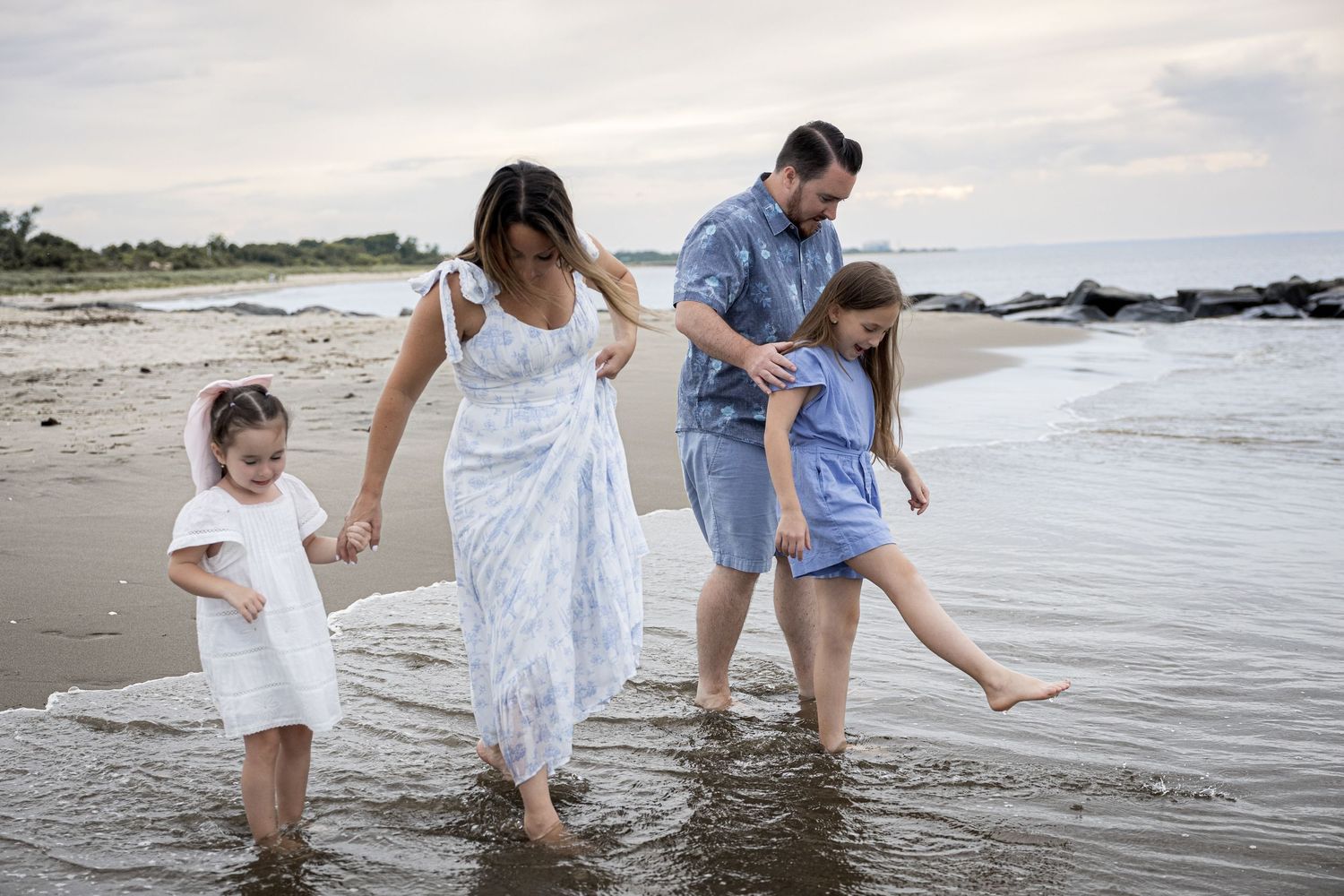 A family of four splashes in shallow ocean water on a cloudy beach day together.