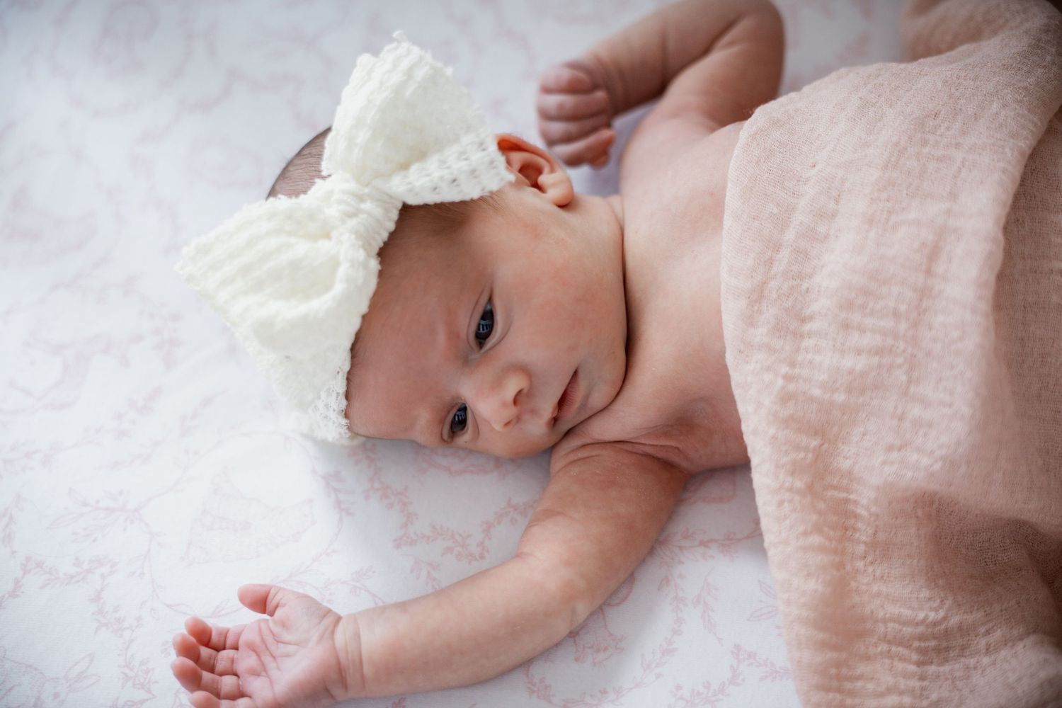 Newborn baby wearing a white bow headband rests on a pink blanket.