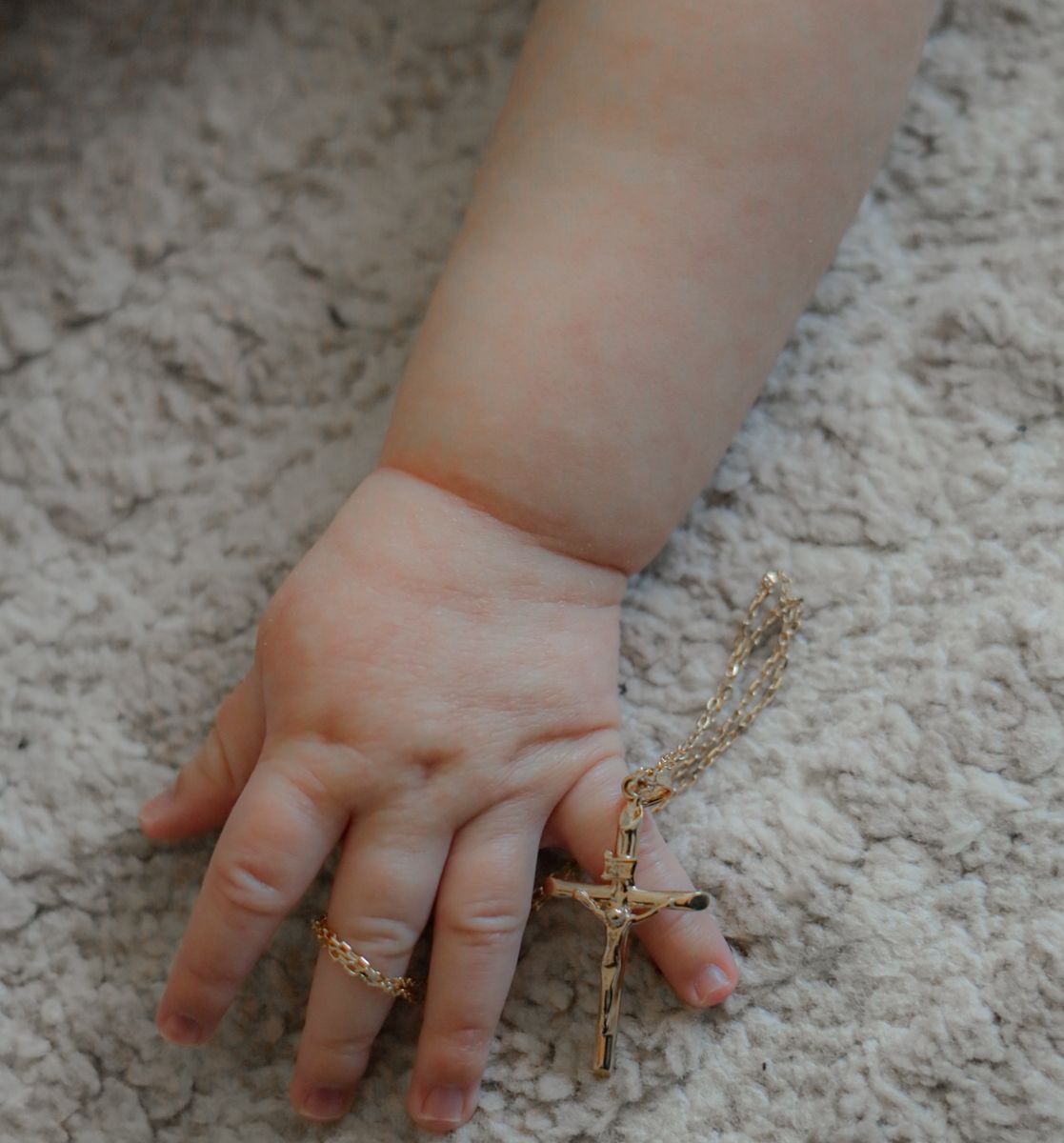 Close-up of a baby's tiny hand gently holding a gold crucifix rosary on a soft carpet.