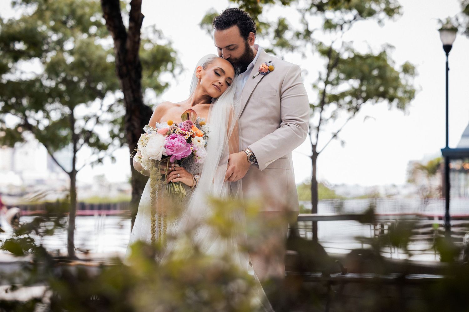 Bride with bouquet leans on bearded groom in beige suit near waterfront with trees in background for New Jersey wedding.