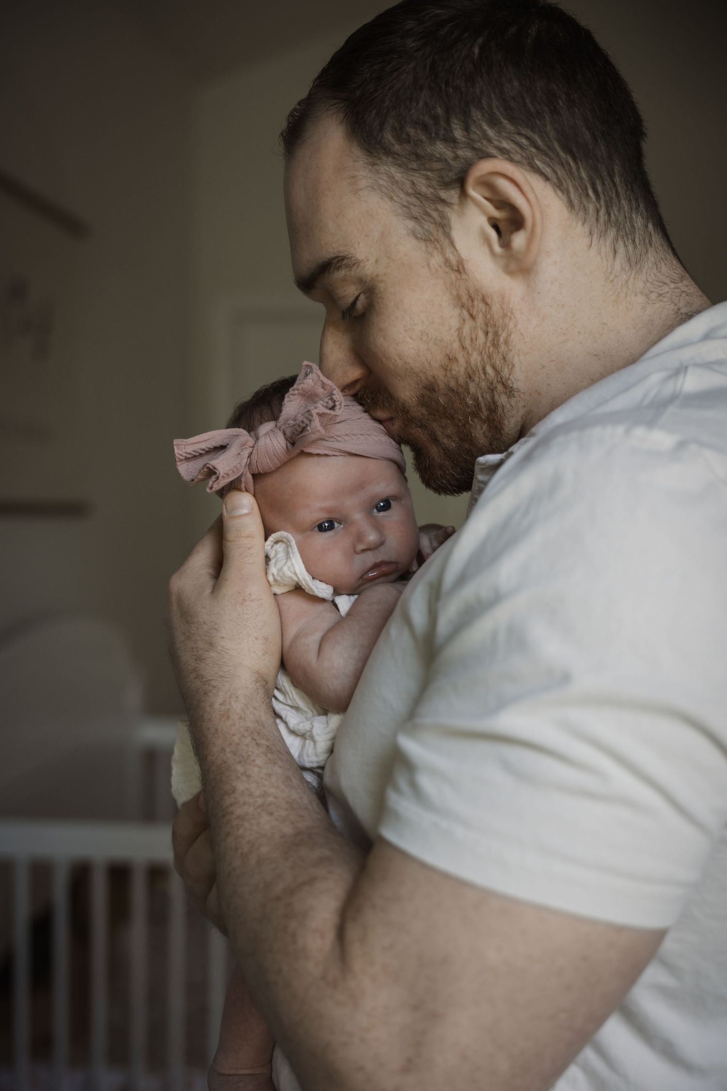 Father tenderly kisses his newborn baby girl wearing a pink bow headband in a nursery.