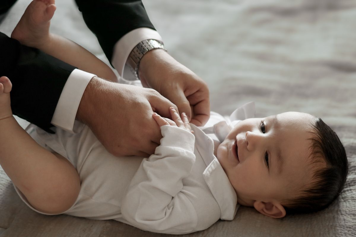Parent in formal suit carefully dressing a peaceful baby in a white onesie on a soft surface.