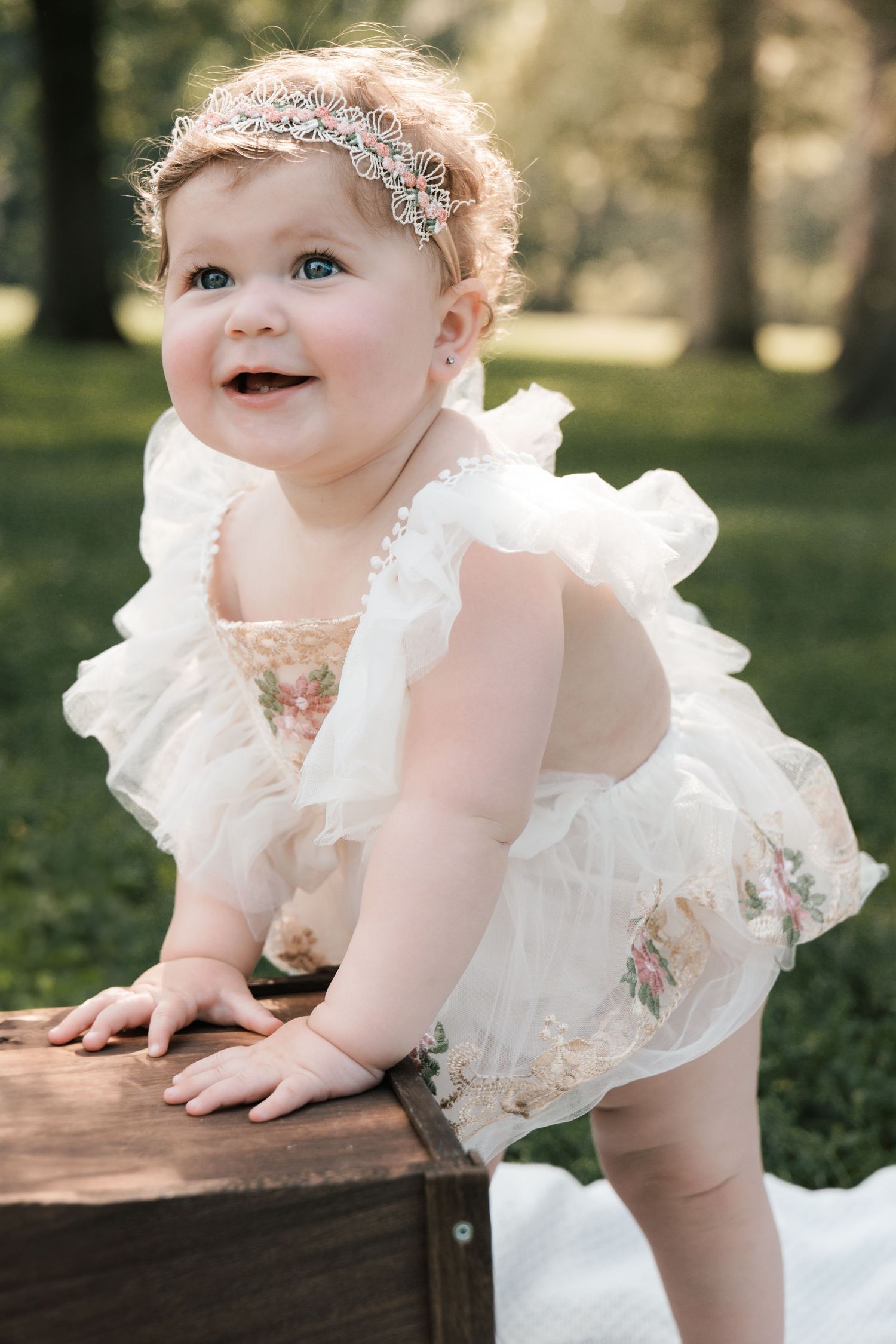 Smiling baby girl in white floral ruffle dress and crystal headband standing outdoors in green park.