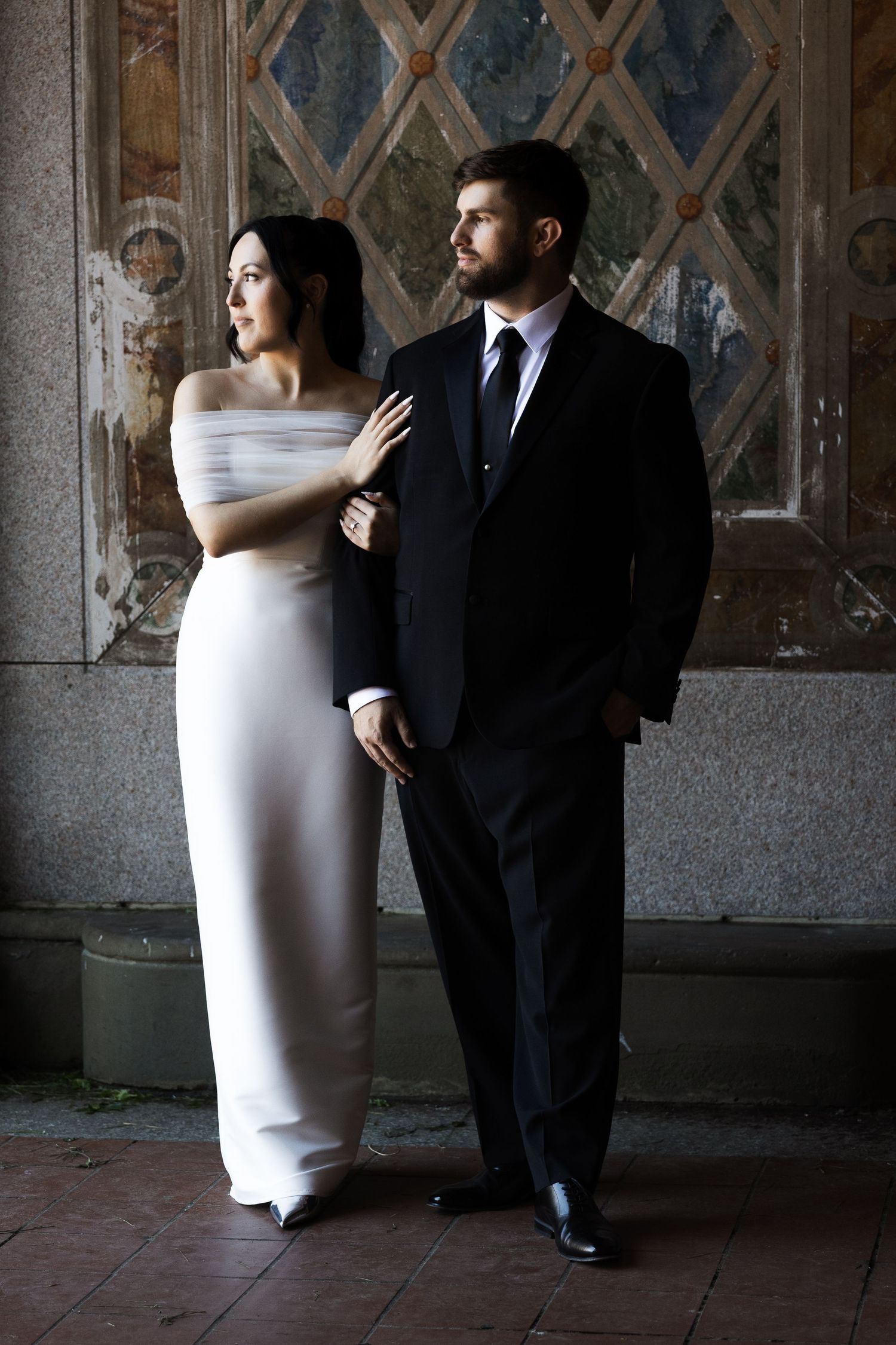 Sophisticated bride in white gown and groom in black suit pose dramatically before ornate stained glass wall in New Jersey.