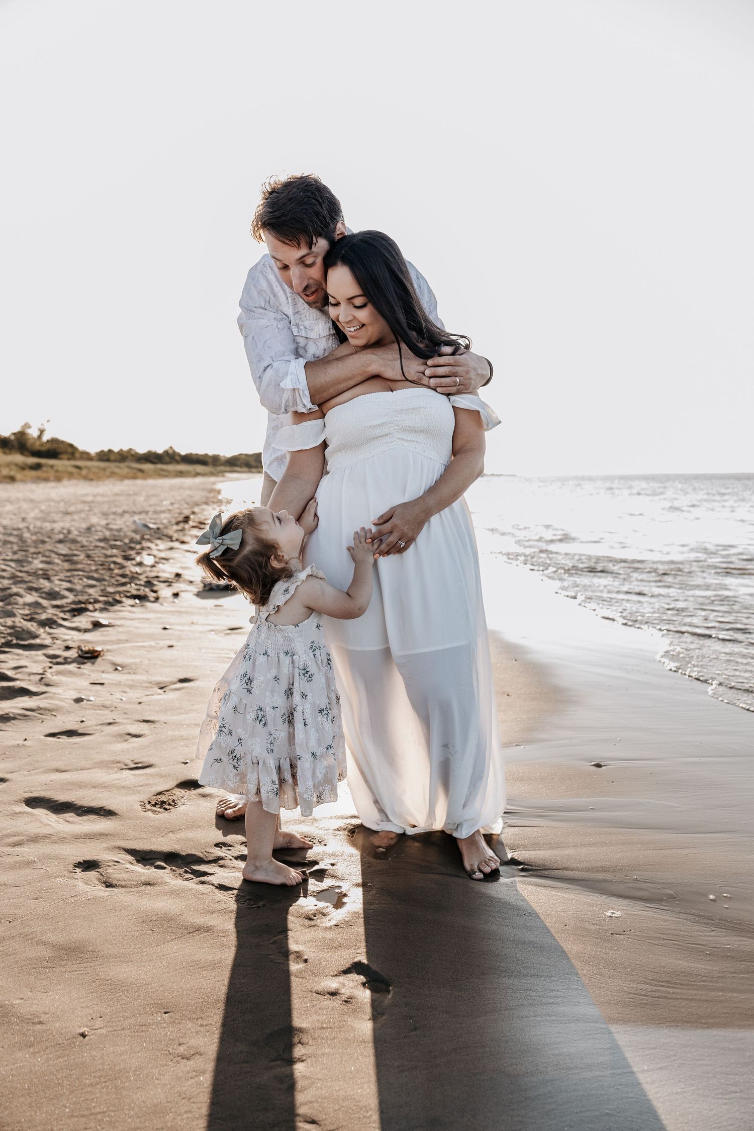 Pregnant woman in white dress embraced by husband and daughter on beach.