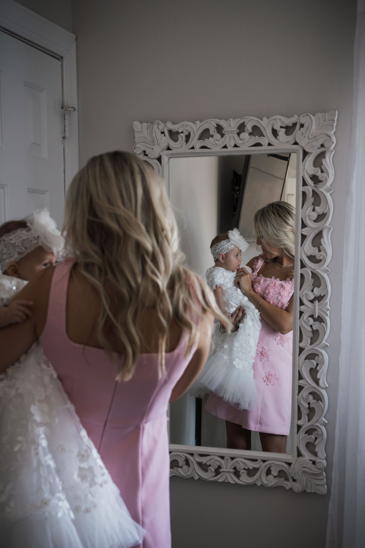 Blonde mother in pink dress holding baby girl in white dress, reflected in ornate white mirror.
