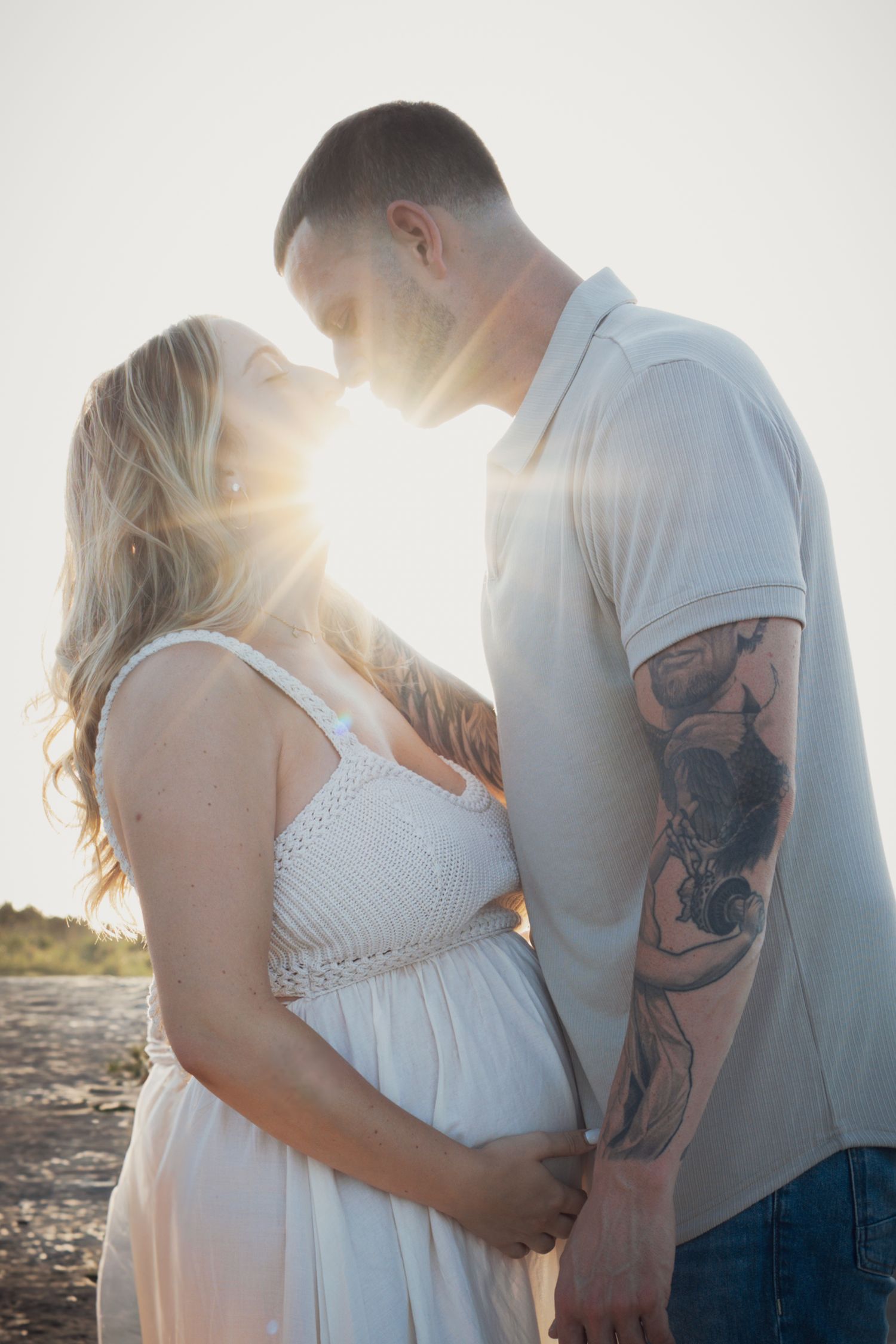 Pregnant couple kissing with sun flare behind them, maternity beach photo.