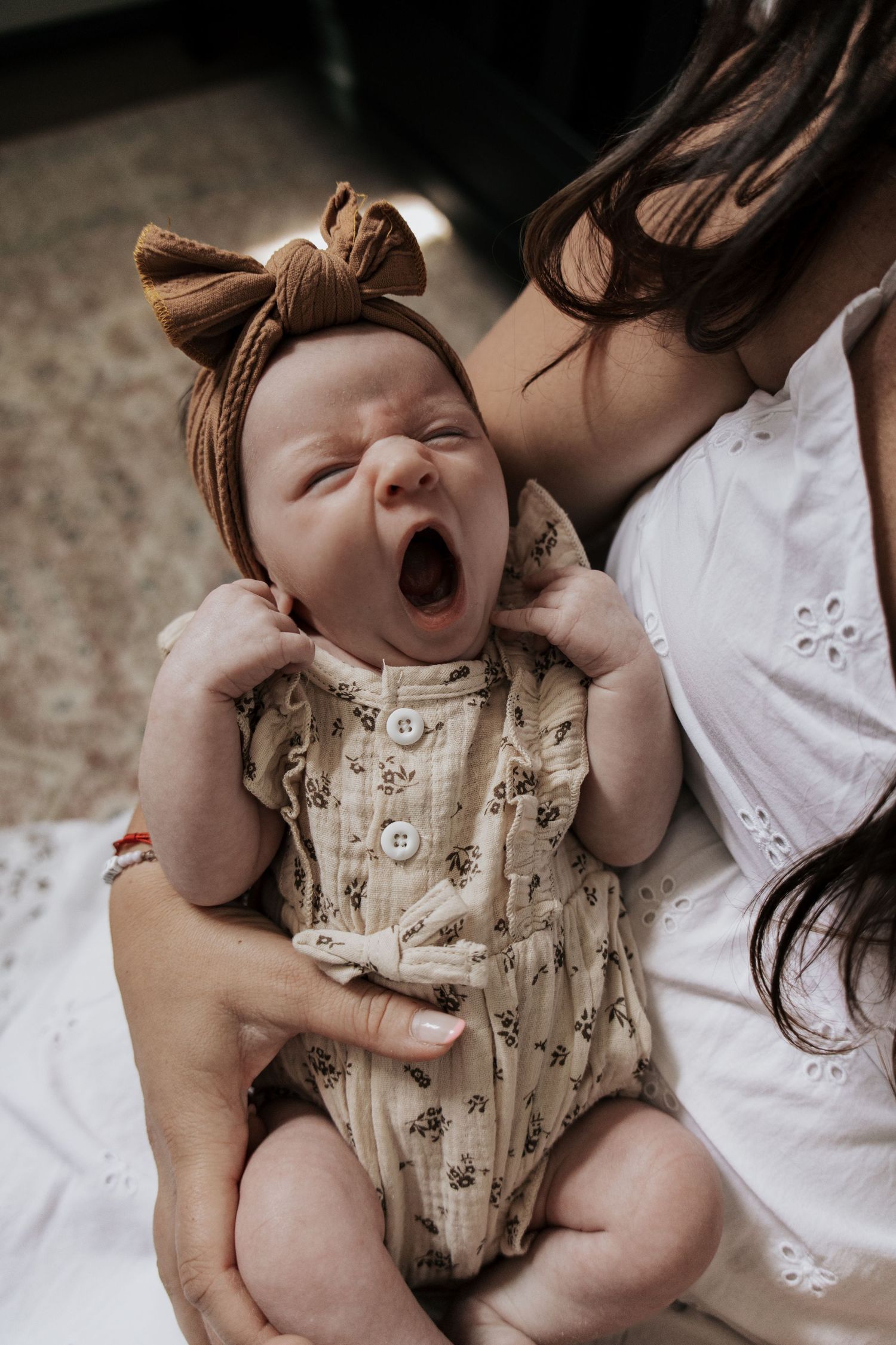 Newborn baby in floral romper and brown bow headband yawning while being held by mother in white dress.