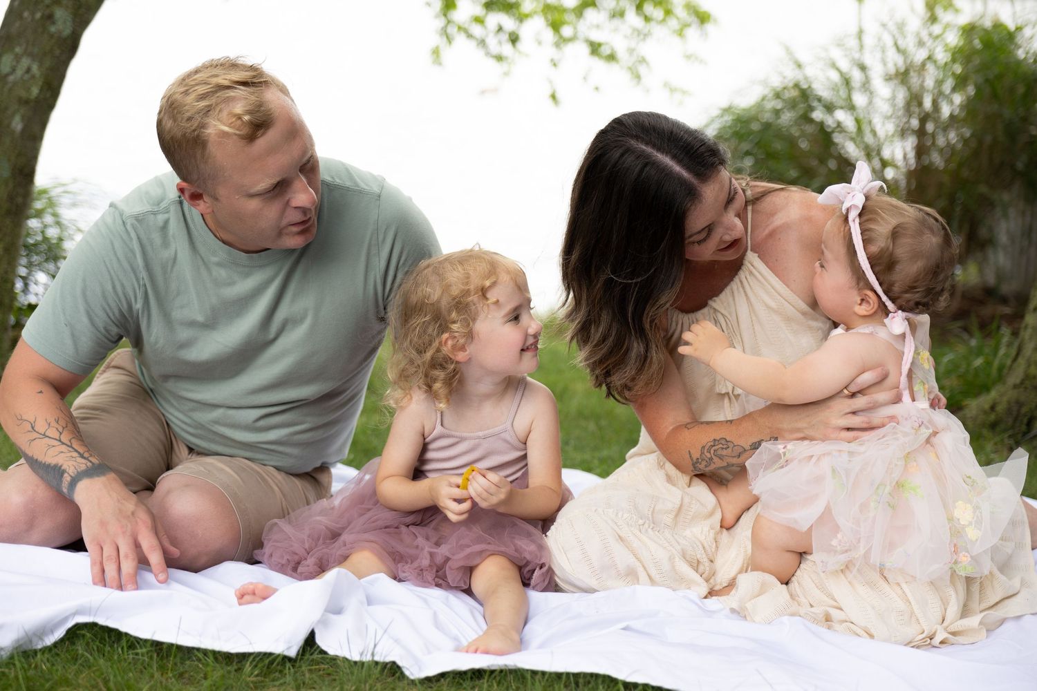 Young family of four sits on blanket in grass near lake celebrating birthday.