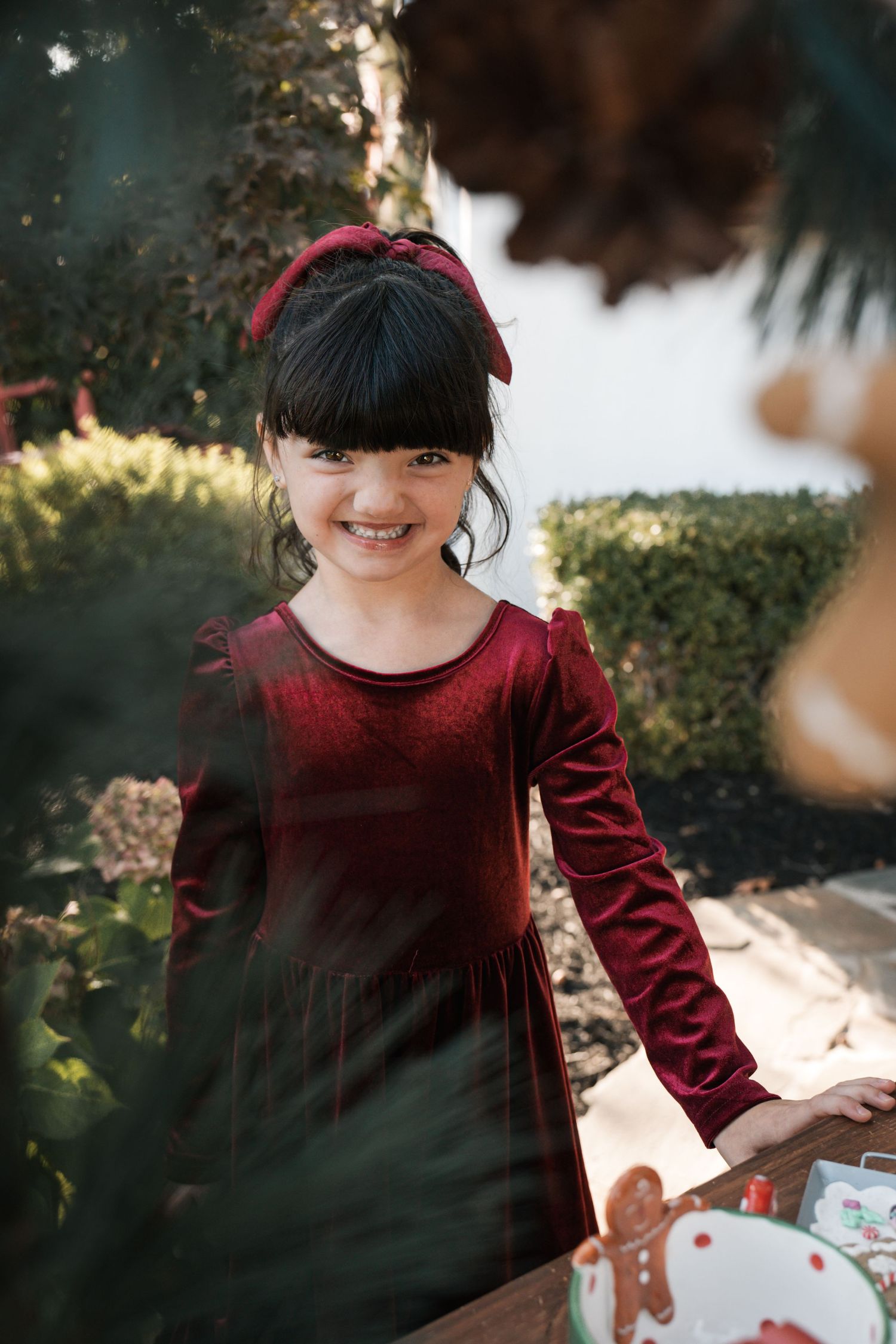 Smiling young girl in red velvet dress with matching bow headband standing outdoors near Christmas decorations.