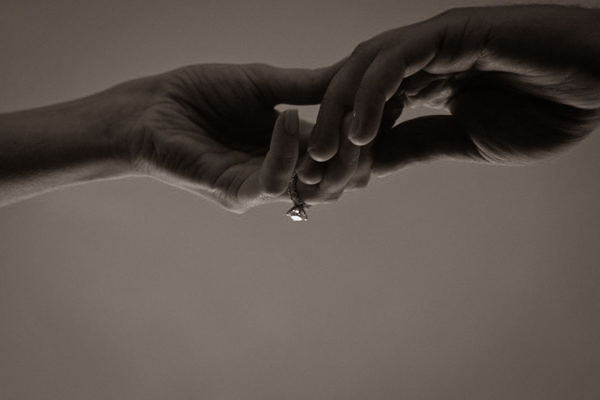 Two hands touching fingertips with a sparkling diamond ring drop between them in sepia tone.