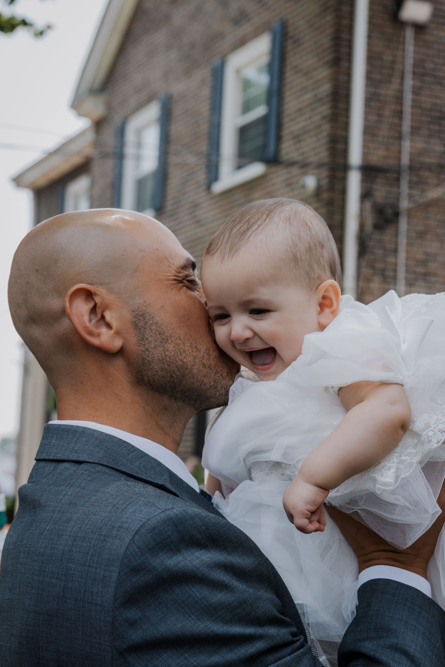 Bald man kissing laughing baby girl in white christening dress outdoors.
