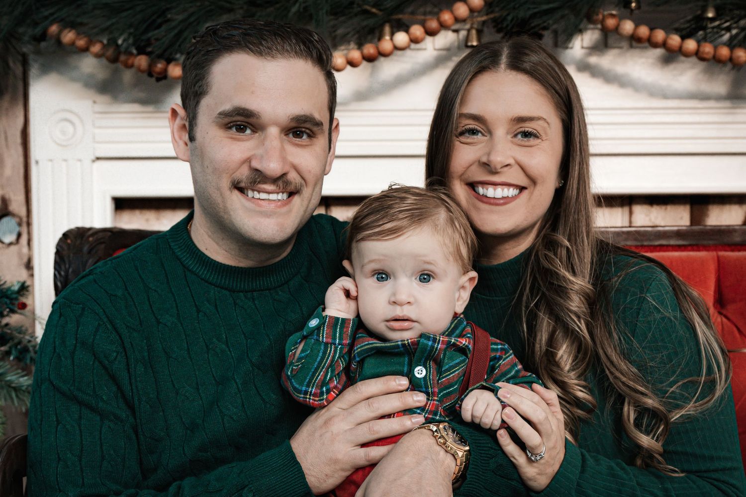 A smiling couple holds their baby boy dressed in plaid for a festive Christmas family portrait.