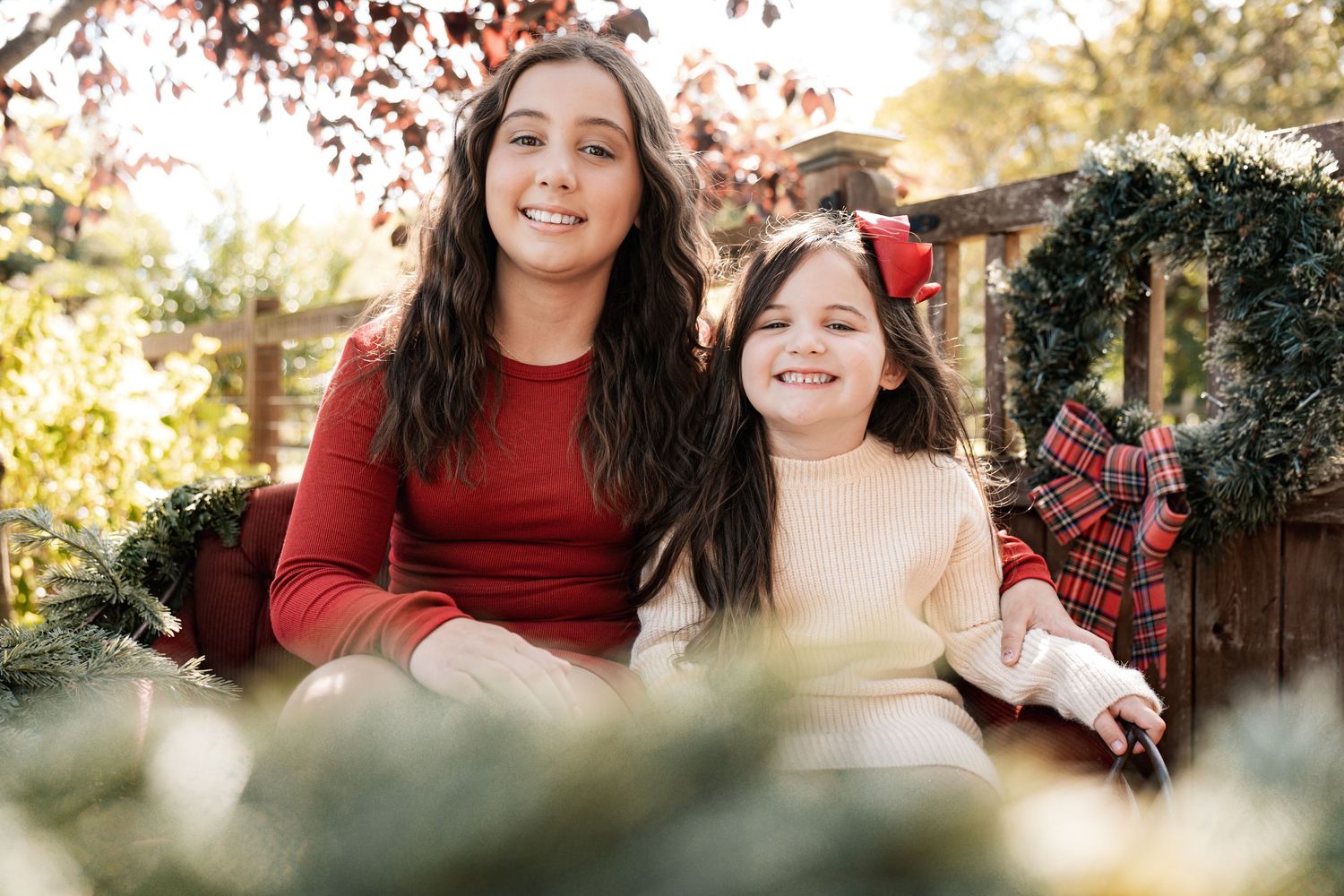 Two sisters smile together on a red velvet outdoor sofa surrounded by festive Christmas wreaths and greenery.