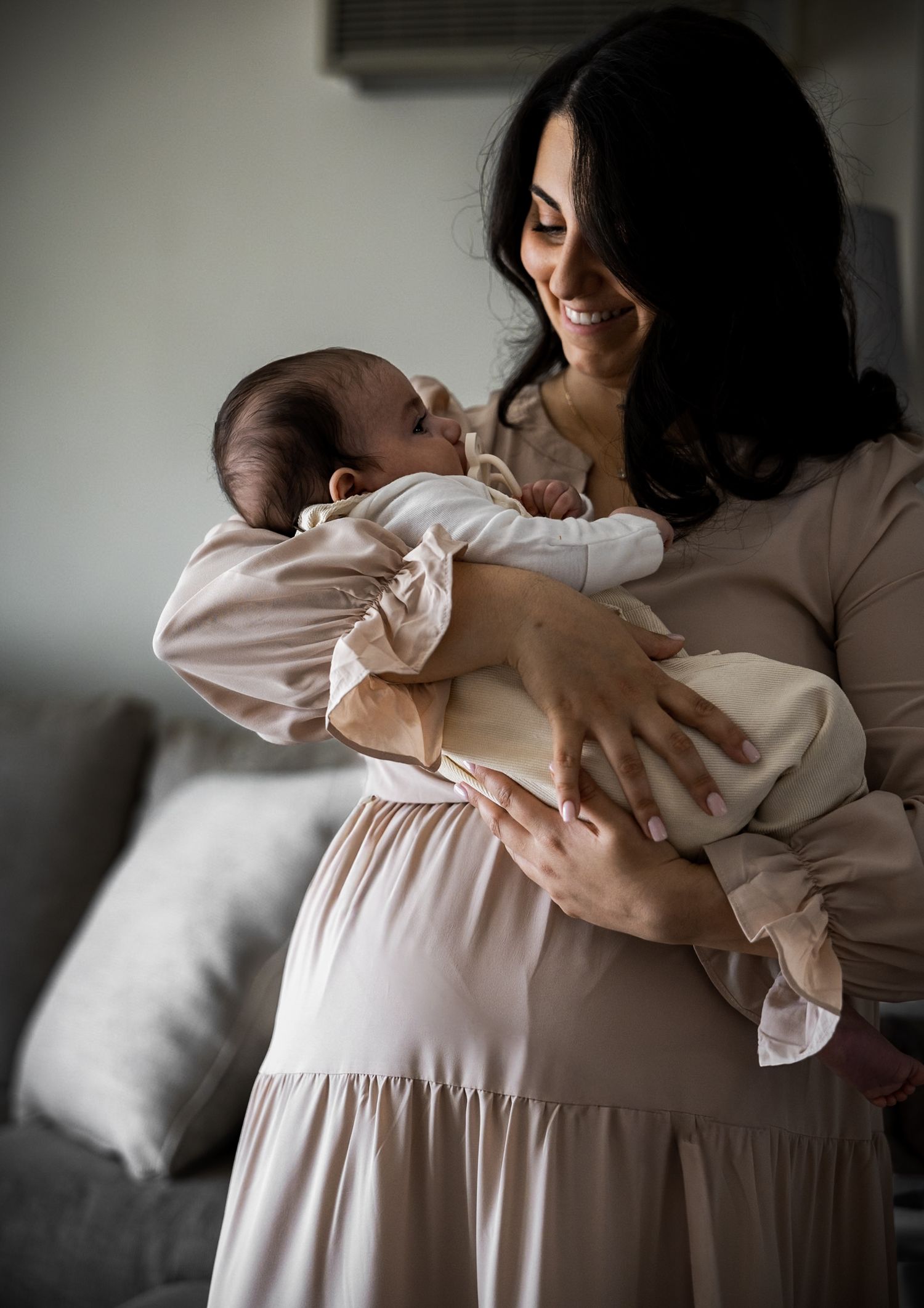 Smiling dark-haired mother in a beige dress lovingly holds her newborn baby.