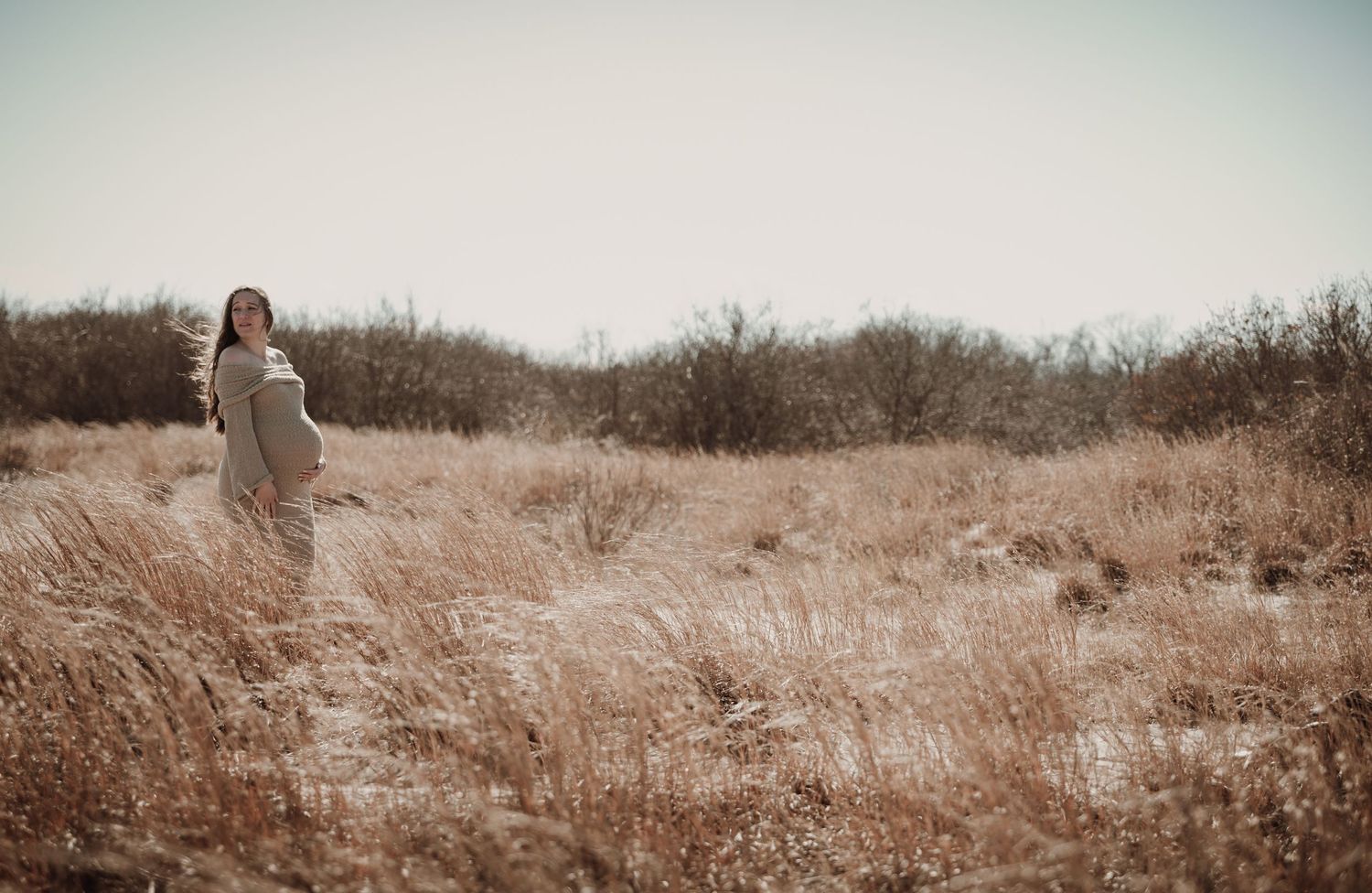 Pregnant woman in tan knit dress standing alone in dry winter field for maternity portrait.