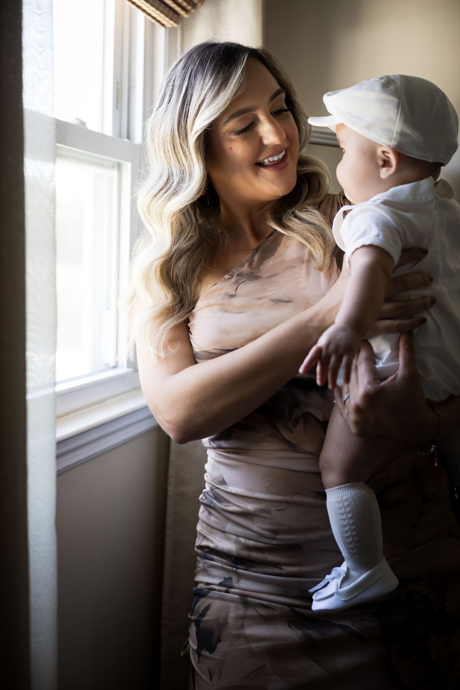 Smiling blonde mother in beige dress holding her baby boy near a sunlit window.