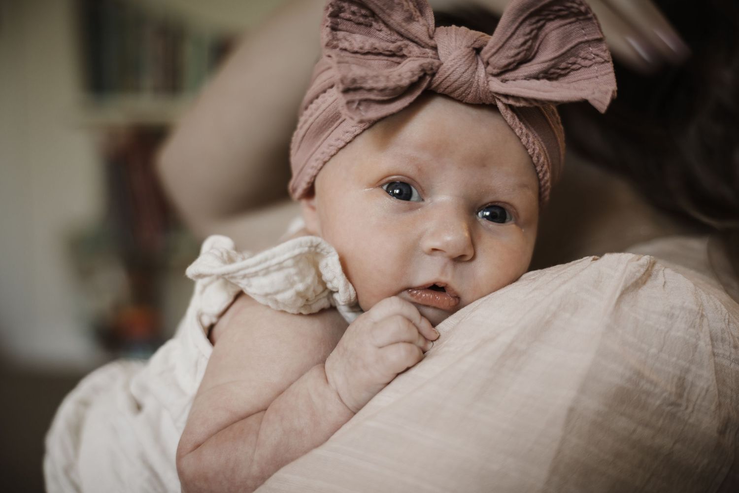 Close-up of a newborn baby girl in a pink bow headband resting on a parent's shoulder.